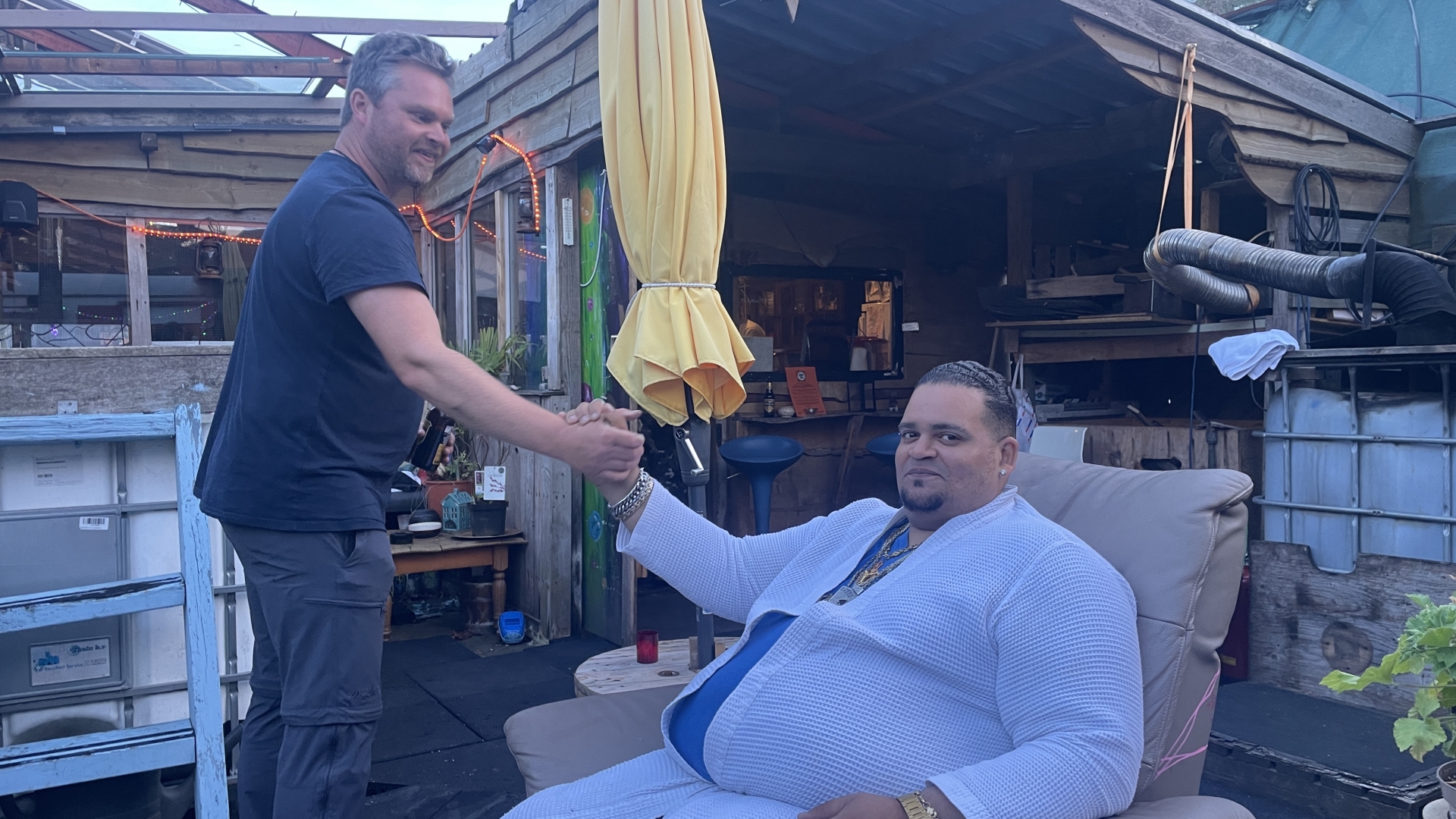 Twee mannen geven elkaar een hand in een buitenruimte met houten muren en een gele parasol.