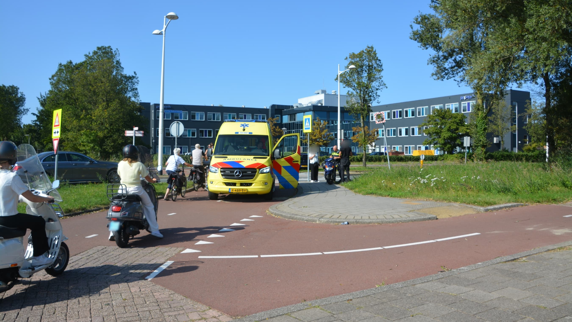 Een ambulance staat geparkeerd op een fietspad bij een kruispunt, met mensen en scooters die om de ambulance heen rijden. Op de achtergrond staan enkele gebouwen en bomen.