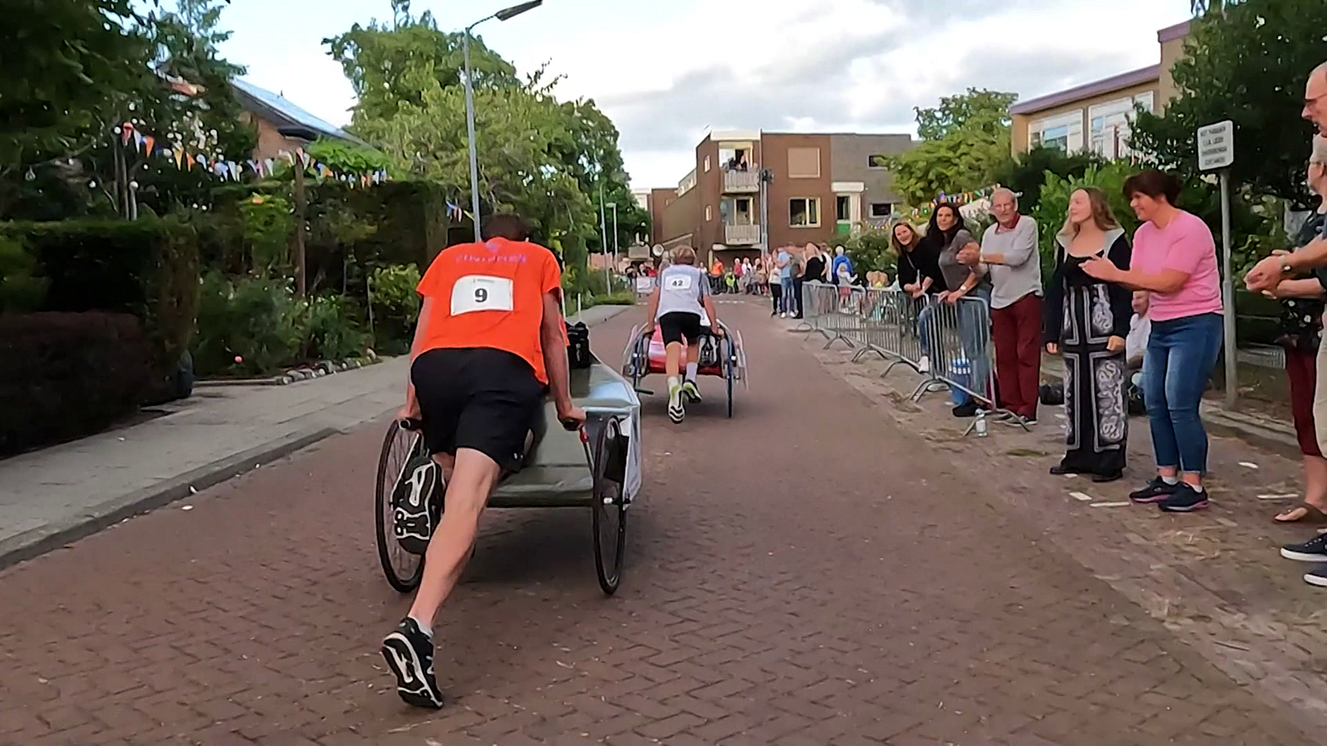 Twee mannen doen mee aan een kruiwagenrace terwijl toeschouwers langs de straat aanmoedigen.