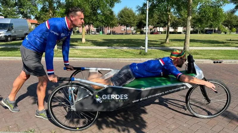 Twee mannen in blauwe sportkleding duwen een speciaal ontworpen fiets op een zonnige dag.