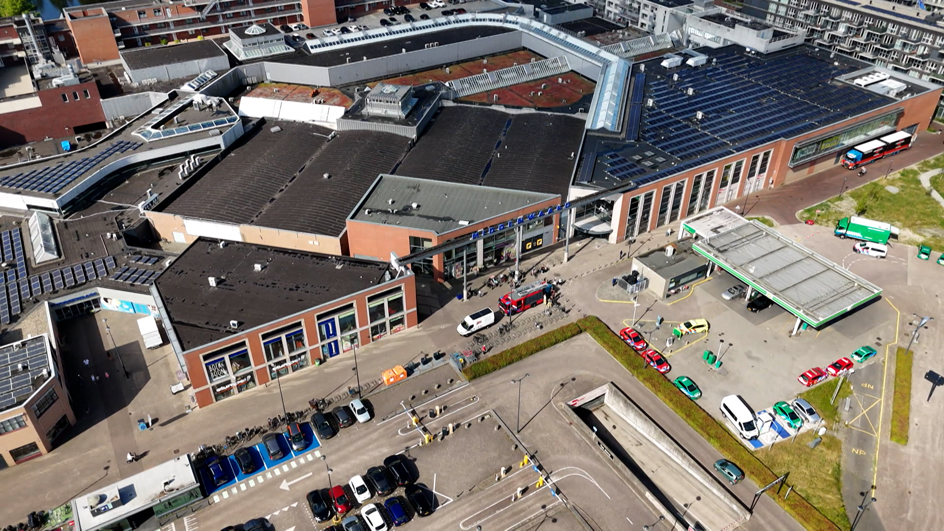 Luchtfoto van een winkelcentrum met zonnepanelen op de daken, parkeerplaatsen, voertuigen en een tankstation aan de rechterkant.