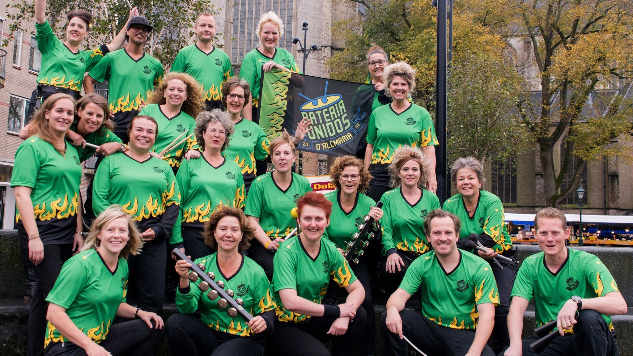 Een groep mensen in groene shirts met vlammenposities, poseren voor een banner met de tekst "Bateria Unidos".