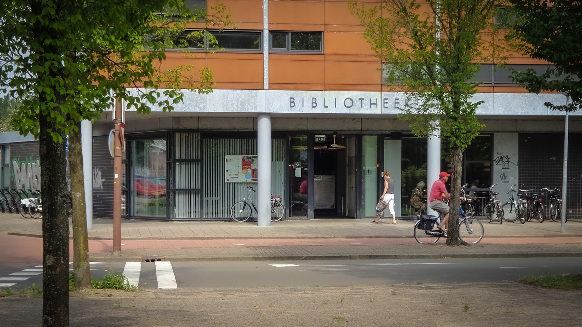 Ingang van een bibliotheek met fietsenrekken ervoor, een fietsende man en een wandelende vrouw.