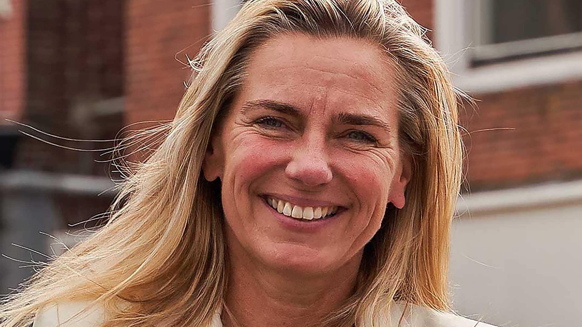 Blonde vrouw glimlachend met een achtergrond van bakstenen gebouwen.