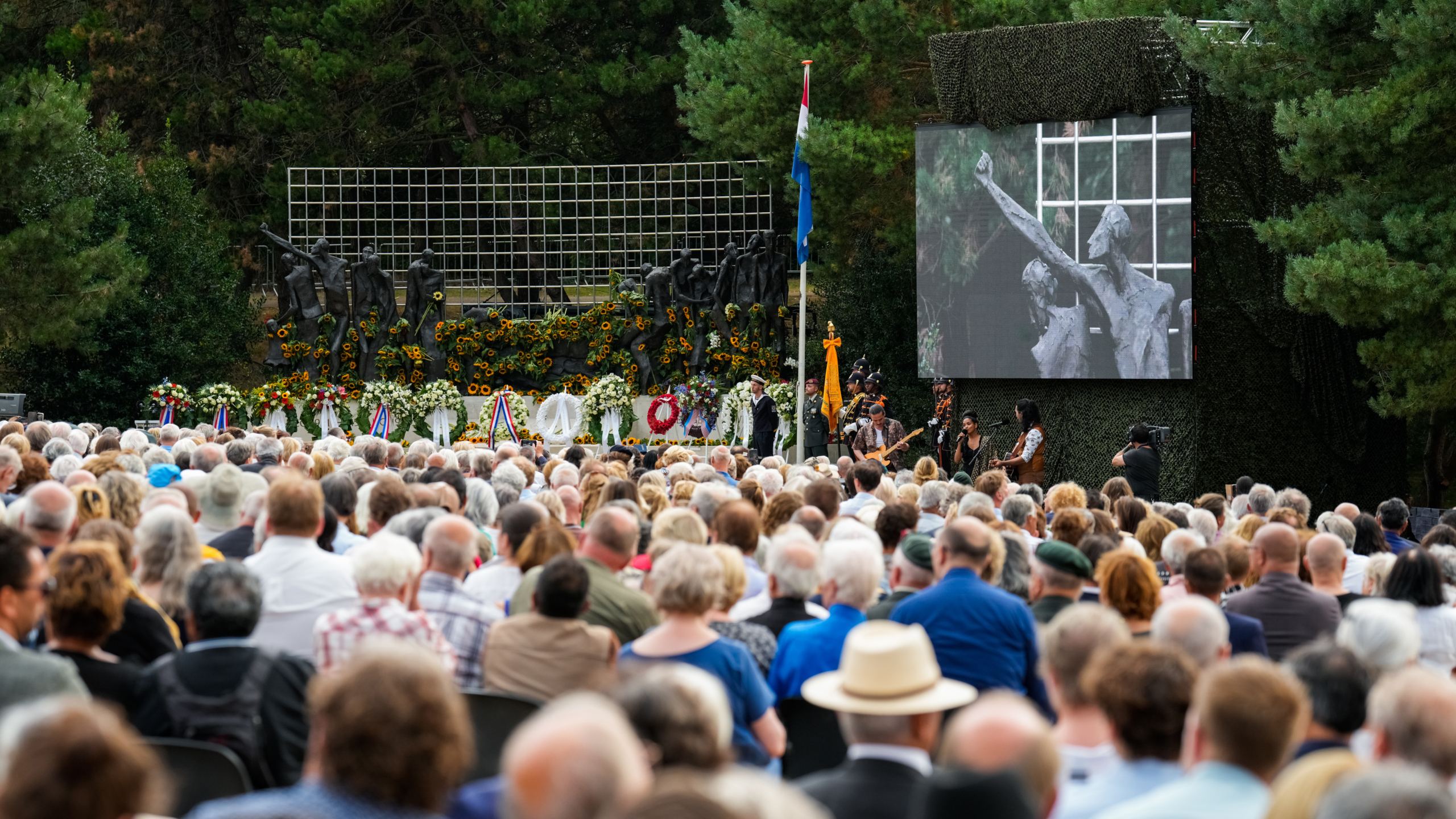 Herdenkingsdienst bij een groot monument met bloemenkransen, veel mensen aanwezig, scherm toont een close-up van het monument.