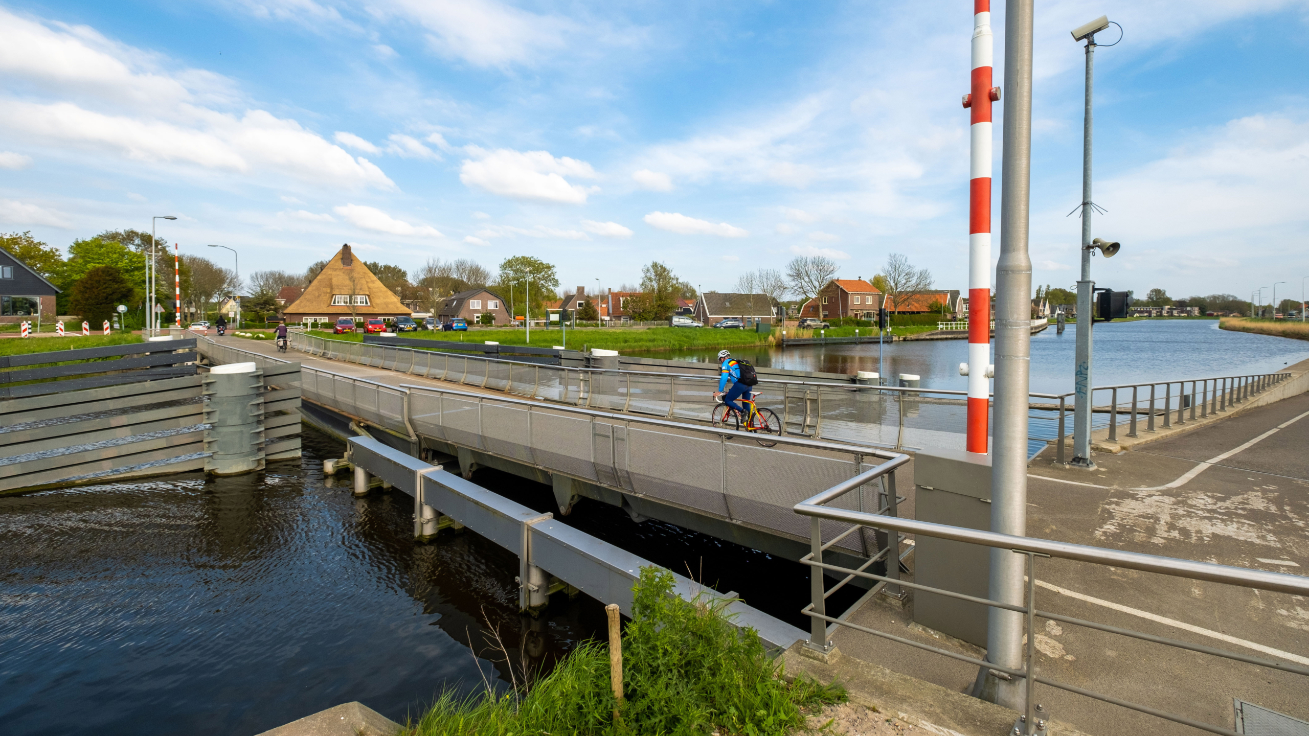 Fietsers rijden over een beweegbare brug over een kanaal met op de achtergrond huizen en een boerderij met een rieten dak.