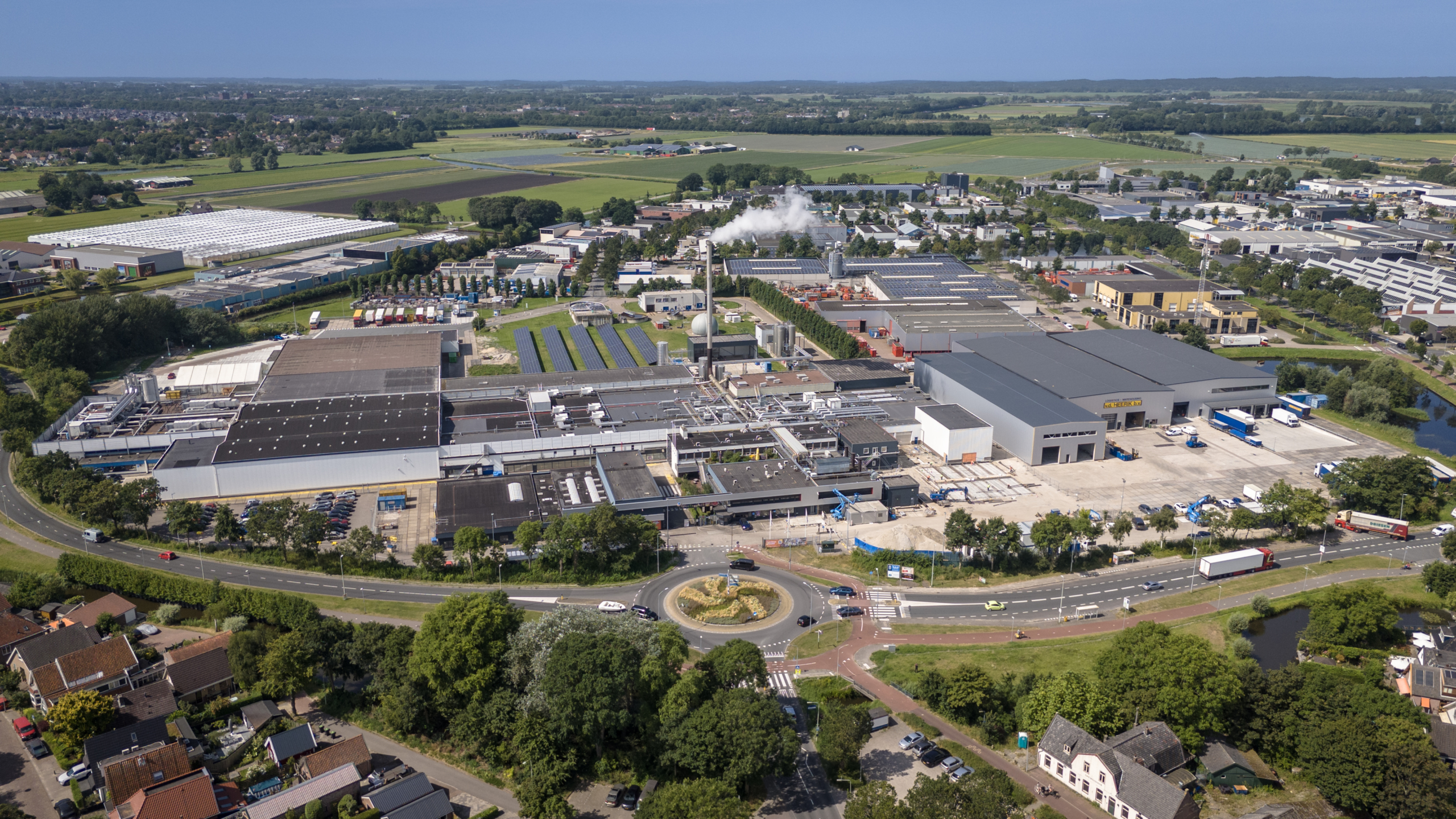 Luchtfoto van een industrieel terrein met grote fabrieken, pakhuizen, vrachtwagens, en omliggende wegen en groen.