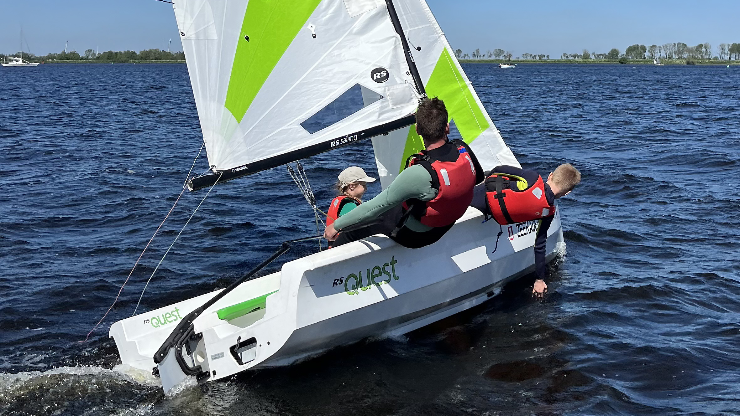 Zeilers in een zeilboot genaamd RS Quest, dragen rode reddingsvesten terwijl ze over het water navigeren.