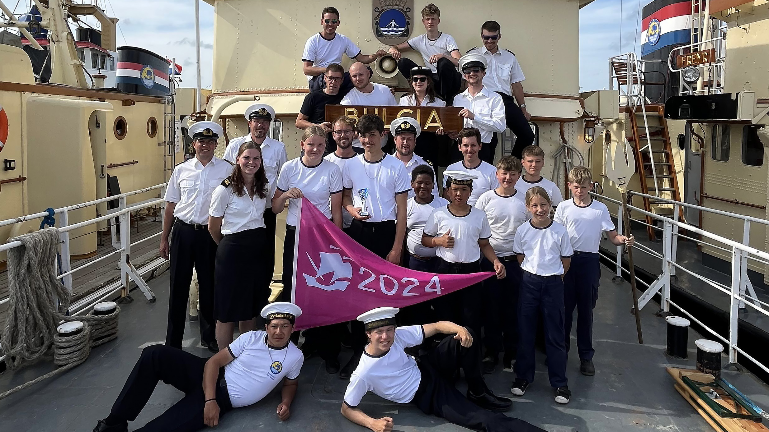 Groep mensen in marine-uniformen poseert op een schip met een roze vlag waarop "2024" staat.