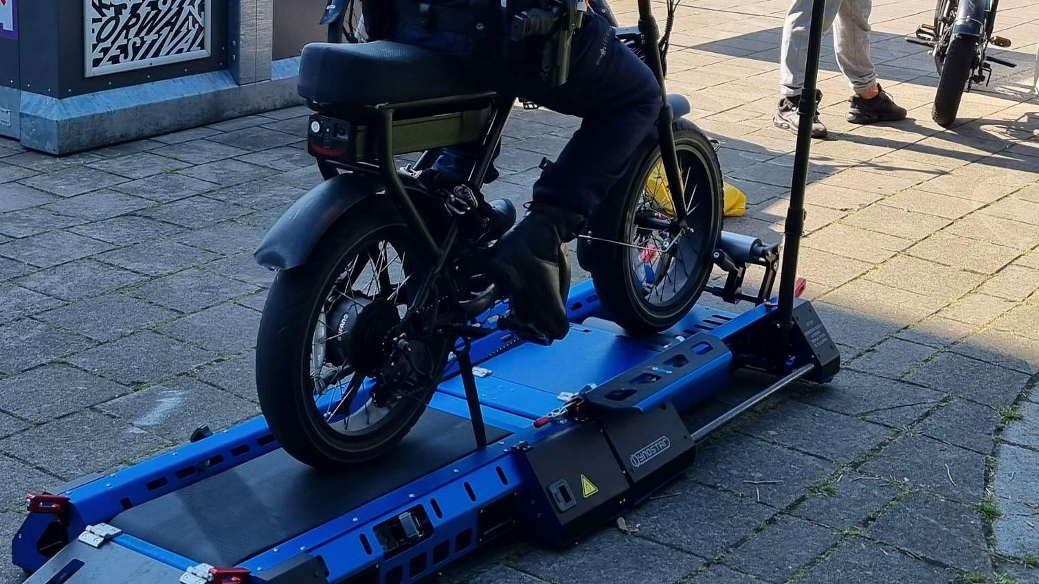 Persoon op een elektrische fiets op een blauwe rollerbank op een trottoir.