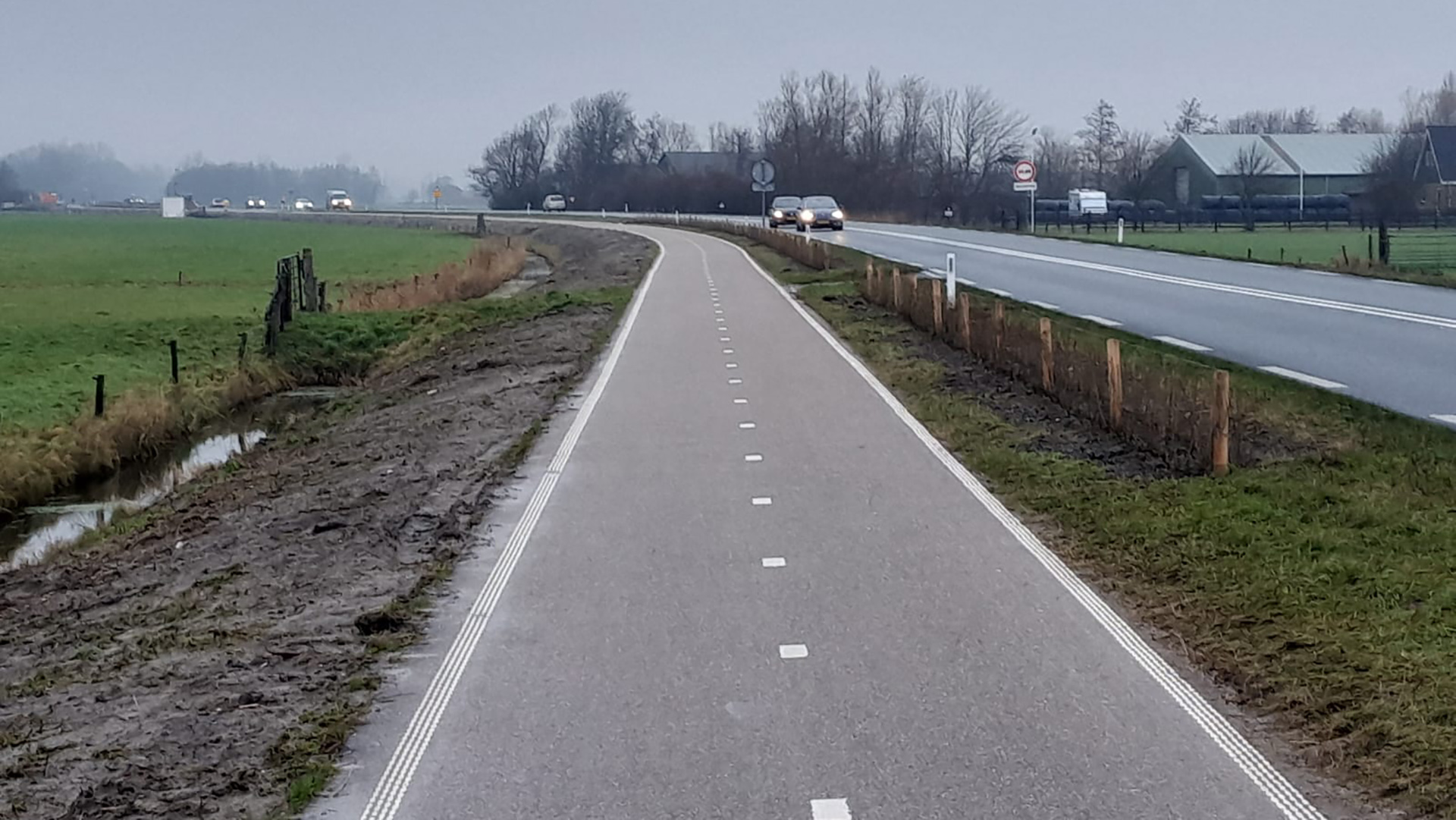 Fietspad naast een weg in een landelijk gebied met grasland en boerderijen op de achtergrond.