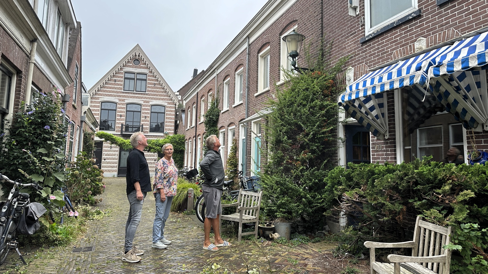 Drie mensen staan in een smalle, met bakstenen omringde straat en kijken omhoog naar een gebouw, met fietsen en groene vegetatie op de achtergrond.