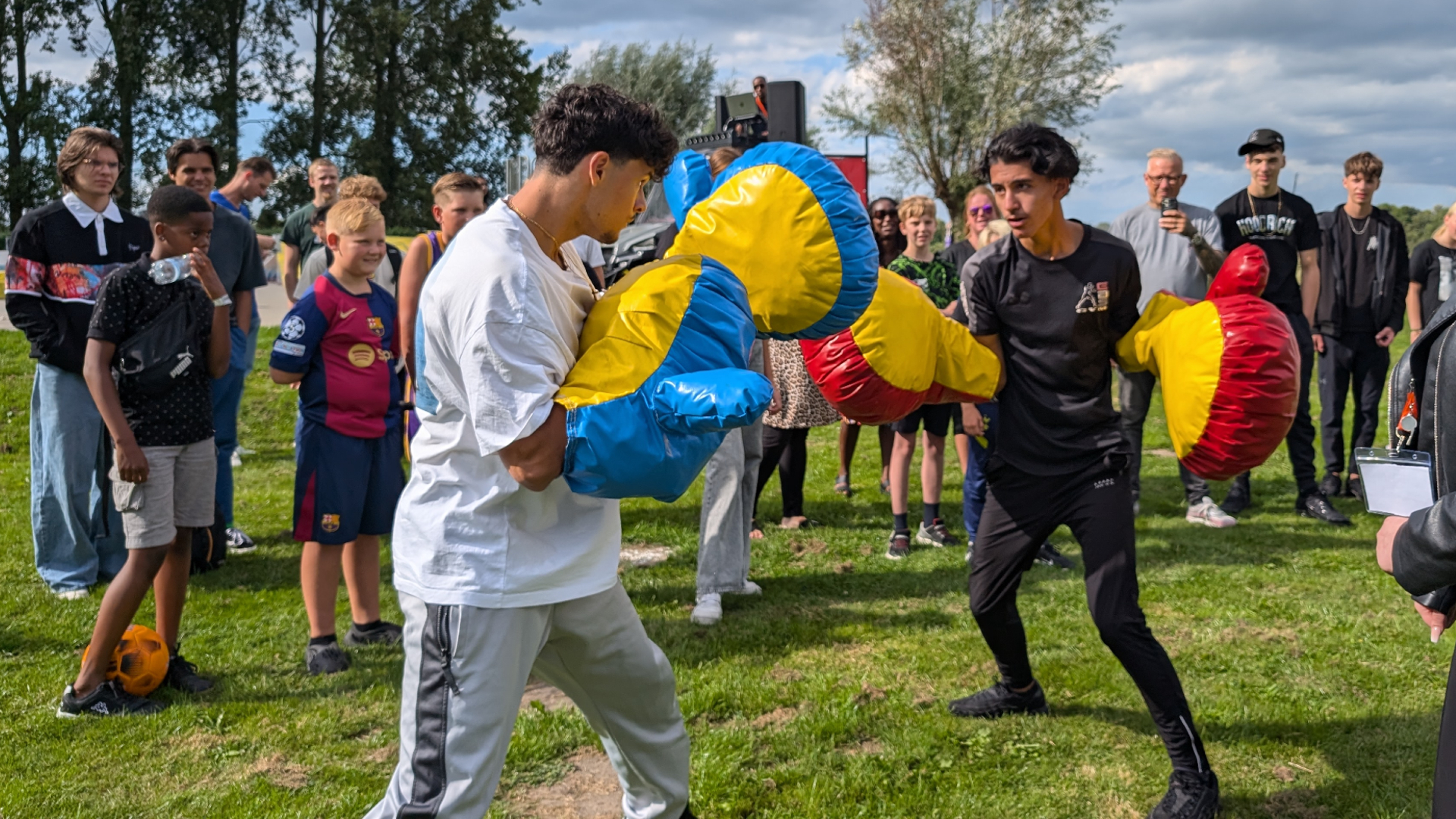 Twee jonge mannen vechten met oversized bokshandschoenen, terwijl een groep mensen om hen heen kijkt op een grasveld.