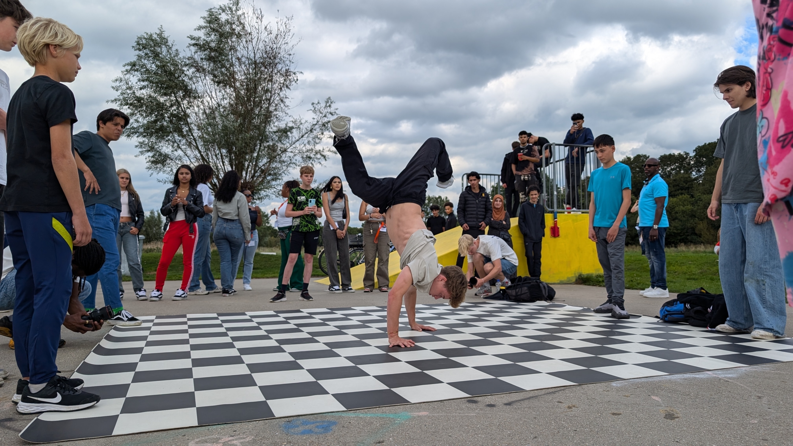 Een tiener voert een breakdance move uit op een zwart-wit geblokte mat, omringd door een groep toeschouwers in een buitenomgeving.