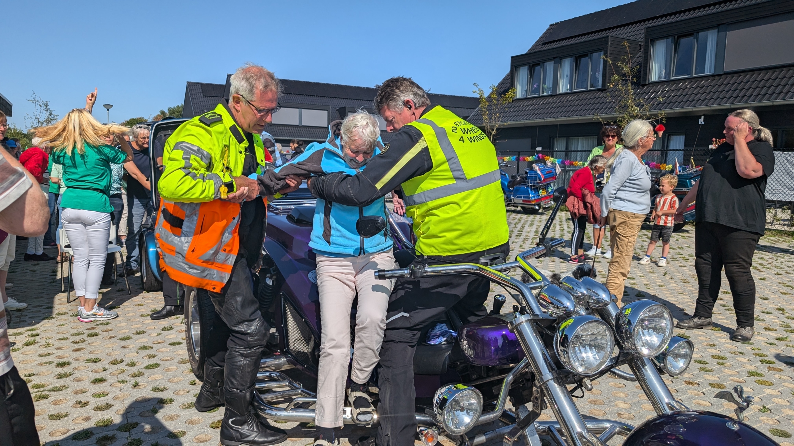 Een oudere vrouw wordt door twee mannen in veiligheidshesjes geholpen bij het afstappen van een driewieler motorfiets, terwijl een groep mensen toekijkt.