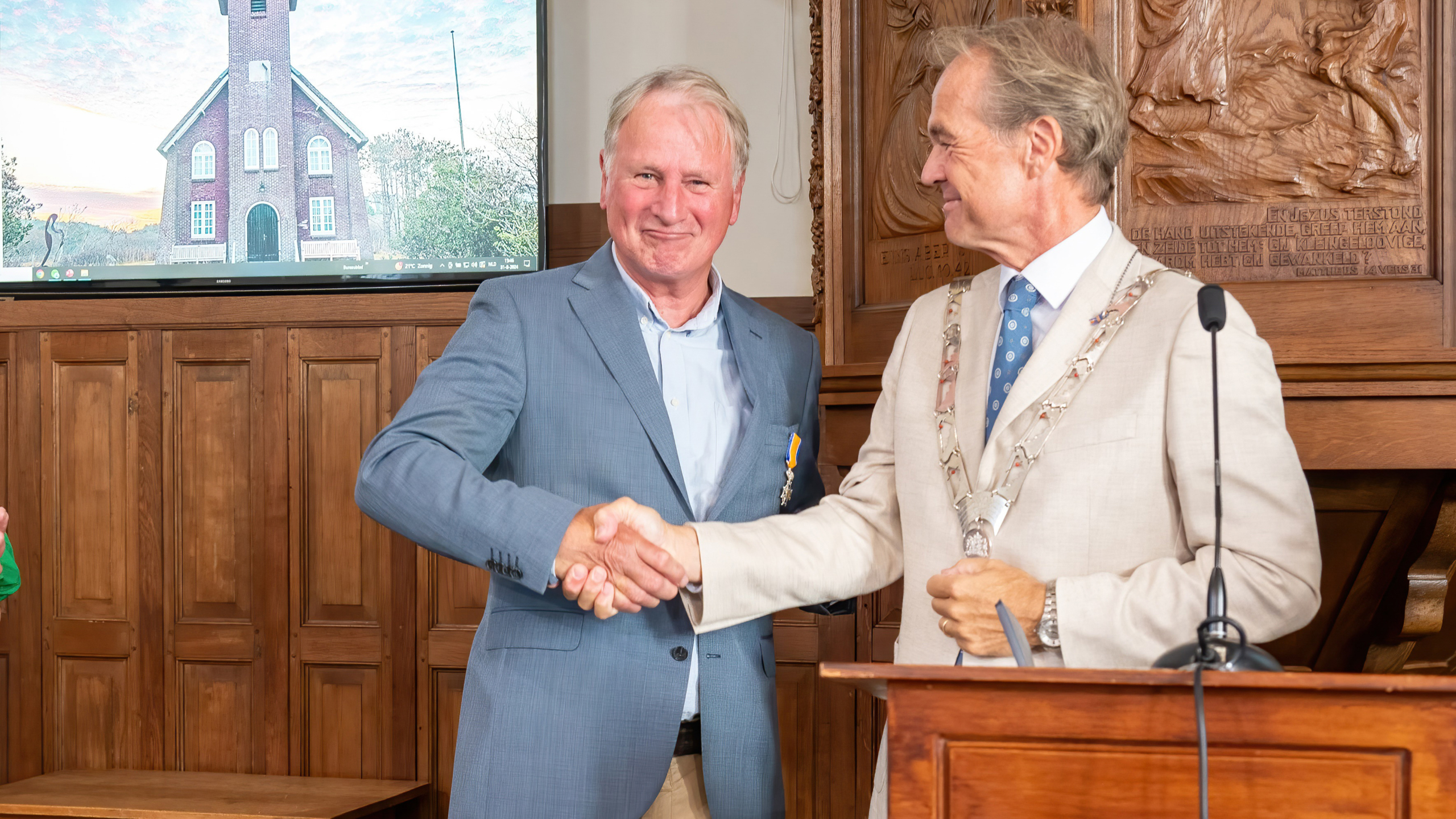 Twee mannen in formele kleding schudden handen tijdens een ceremonie in een houten kamer; één man draagt een lintje en de andere een ketting met medailles.