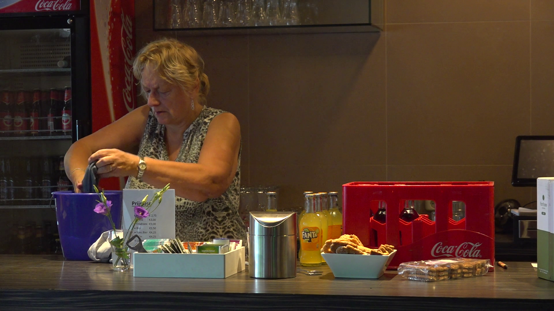 Vrouw werkt achter een bar met frisdrankflesjes en snacks op de toonbank.