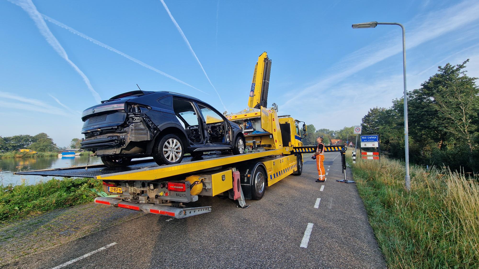 Autowrak wordt op een bergingswagen geladen naast een weg met een waterlichaam en een verkeersbord met de tekst "West-Graftdijk".