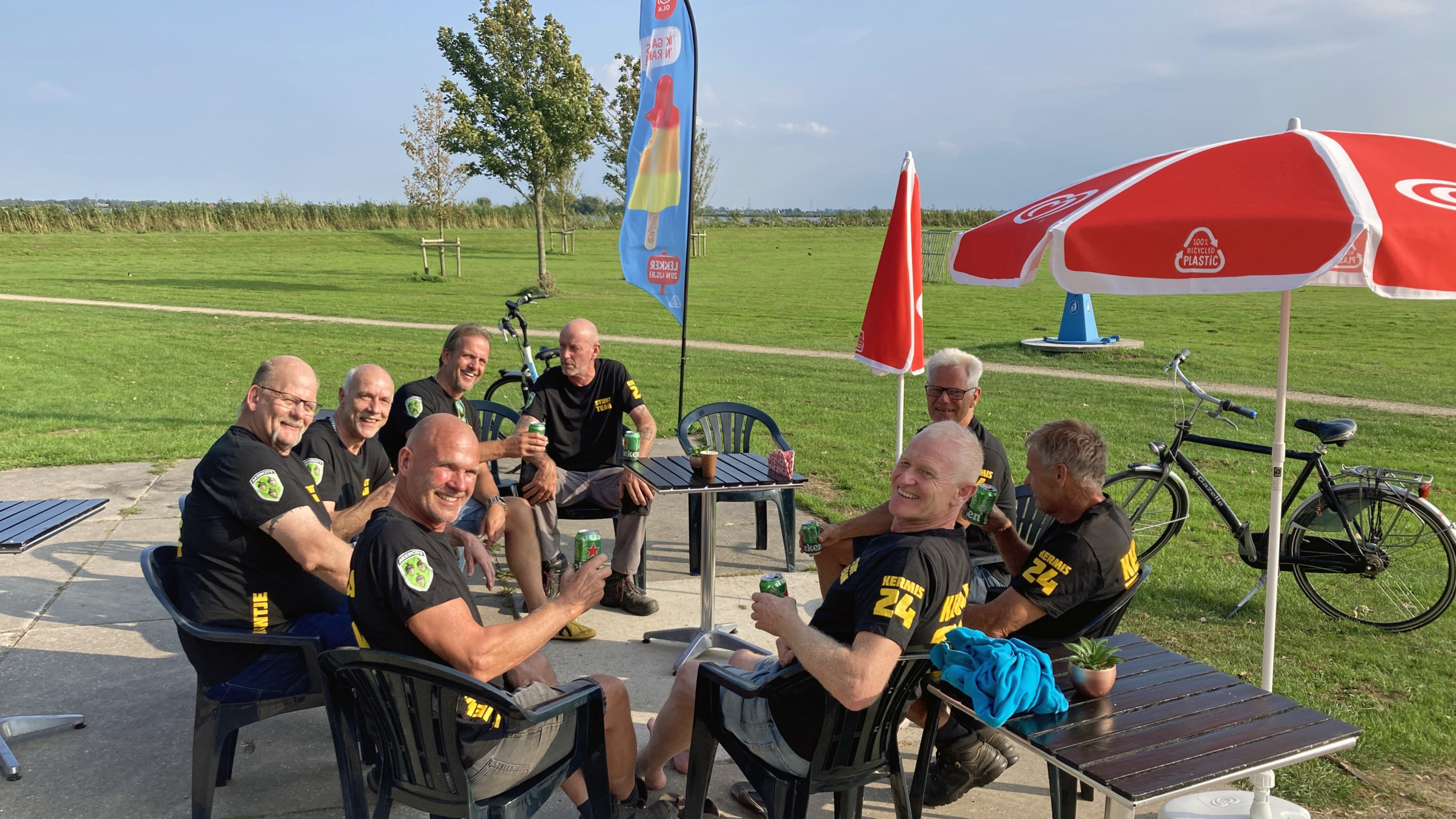 Groep oudere mannen in zwarte shirts met bier, zittend buiten aan tafels met rode parasols en fietsen op de achtergrond.