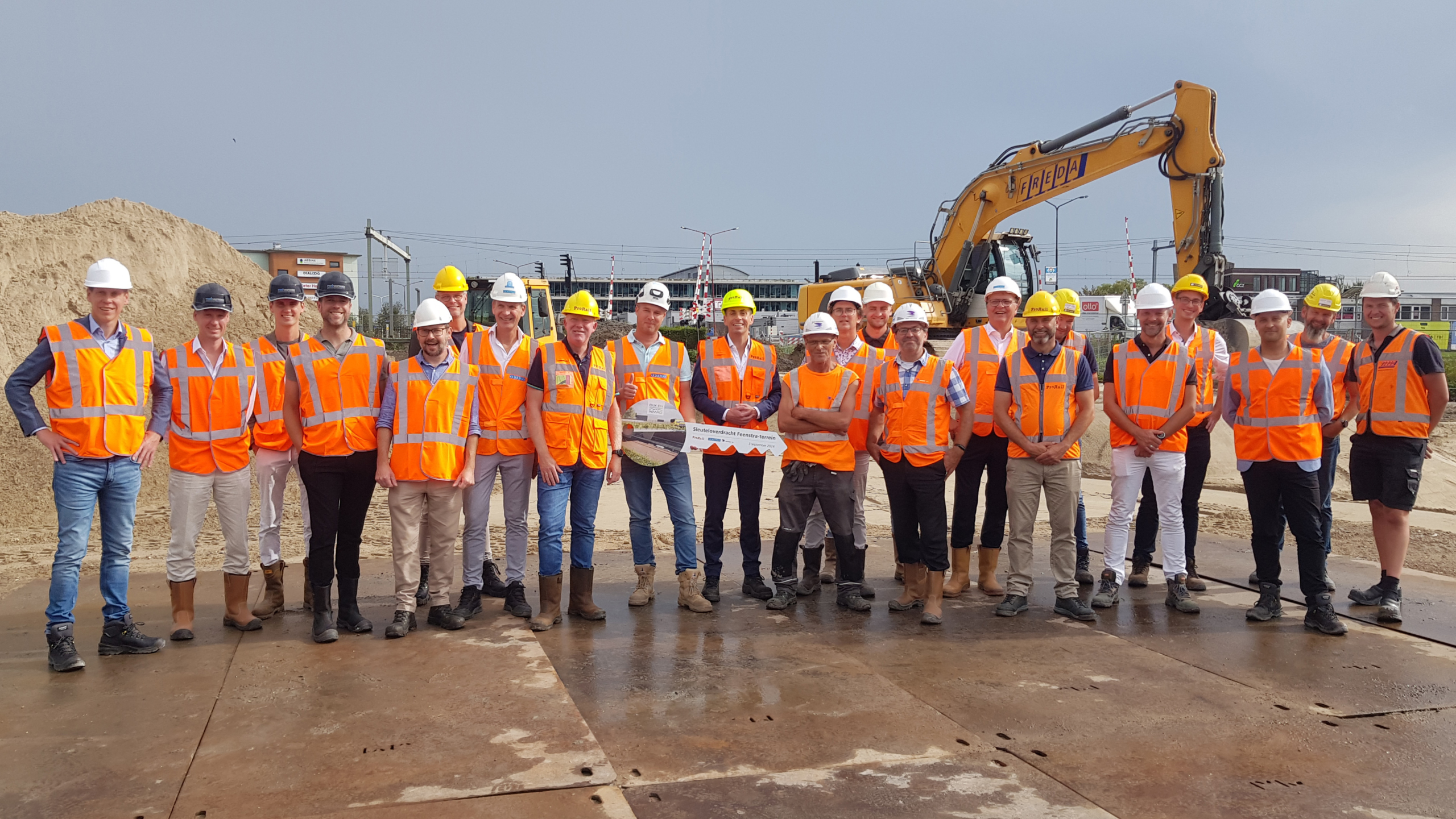 Groepsfoto van bouwvakkers in oranje hesjes en helmen op een bouwplaats met een graafmachine op de achtergrond.