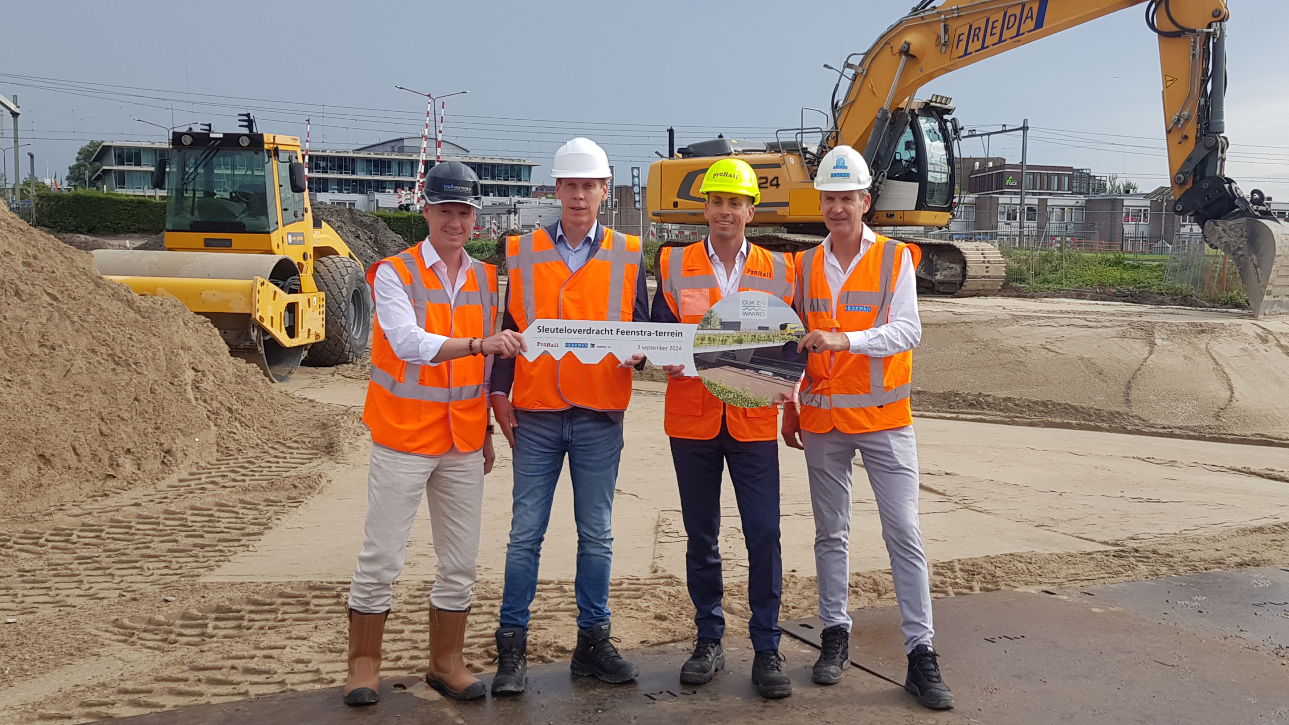 Vier mannen in oranje veiligheidshesjes en witte helmen poseren op een bouwplaats met een bord “Sleuteloverdracht Feenstra-terrein”.