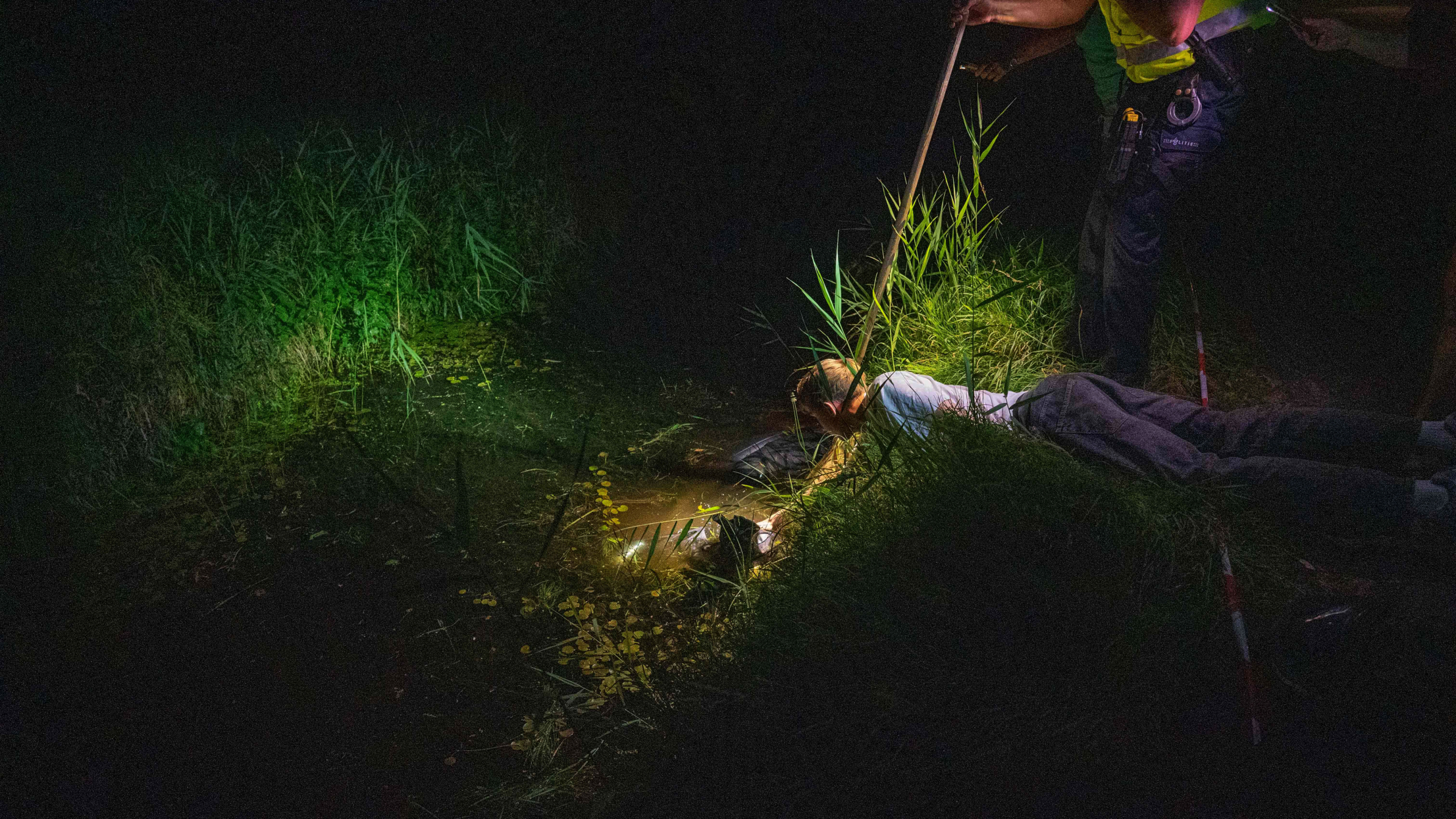 Een persoon ligt op de grond bij een plas water, terwijl een andere persoon met een stok helpt in een donkere, beboste omgeving.