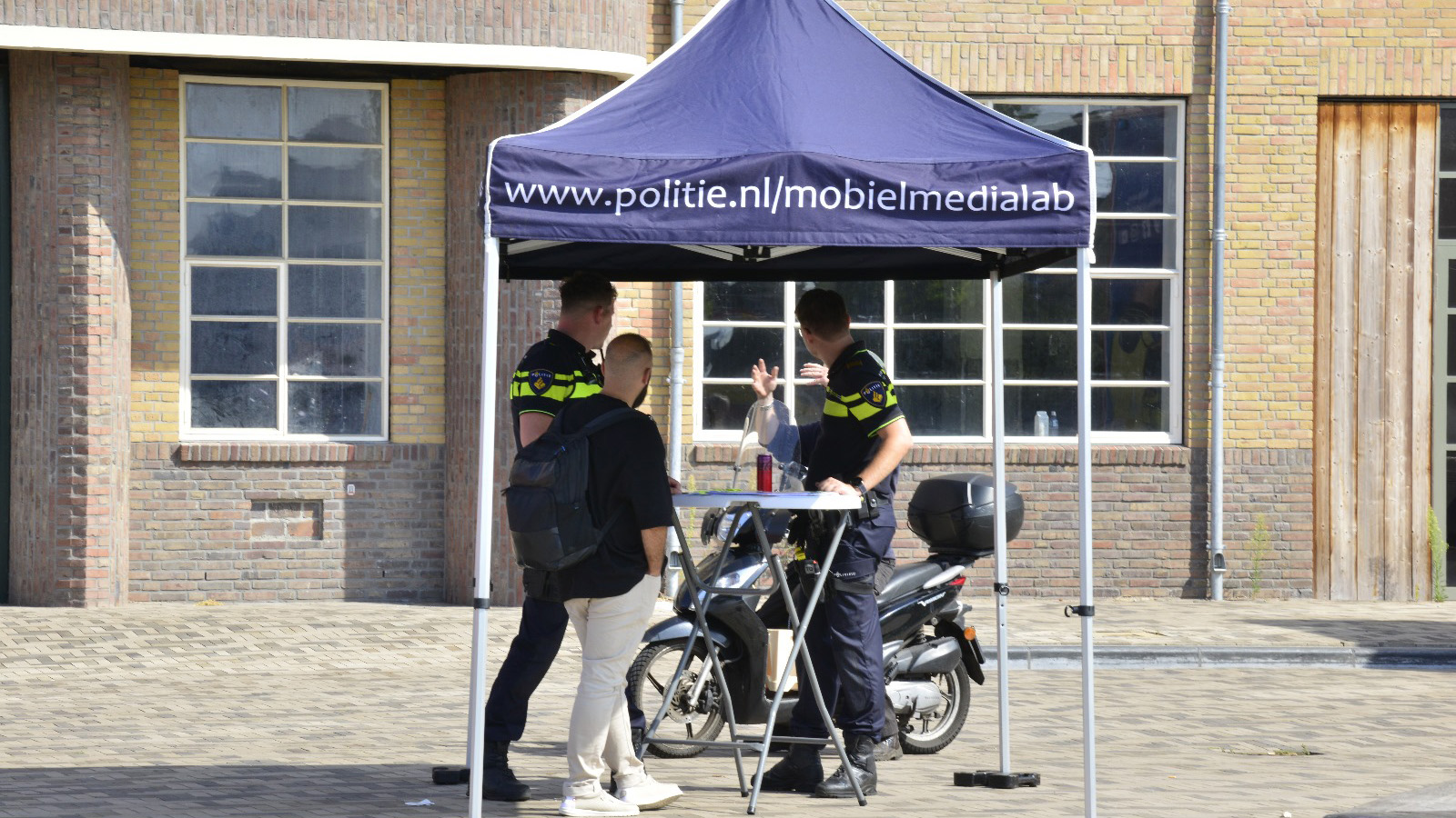 Politieagenten staan onder een tent van politie.nl/mobielmedialab en praten met een man.