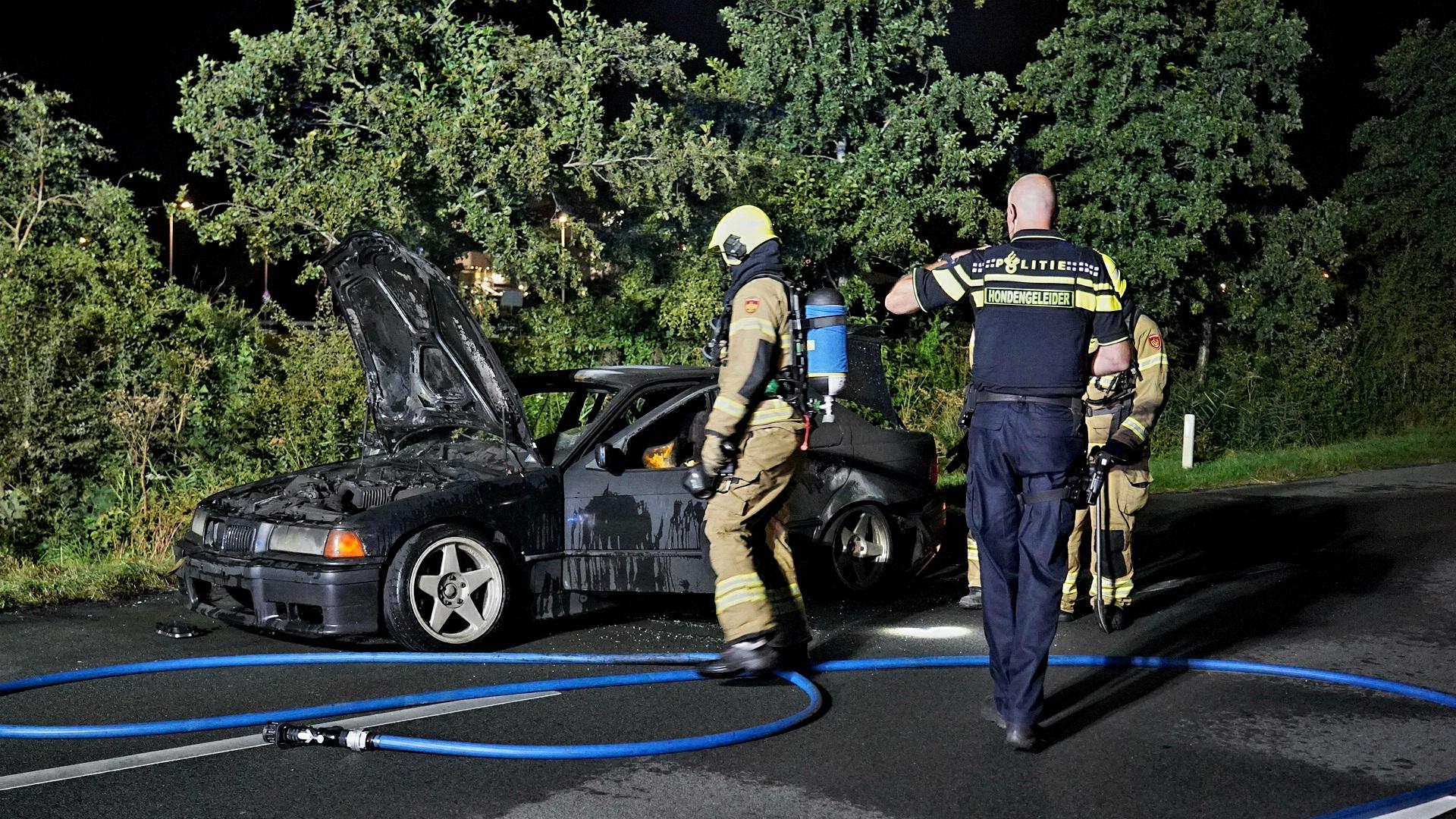 Brandweerlieden en een politieagent onderzoeken een uitgebrande auto langs de weg in de nacht.