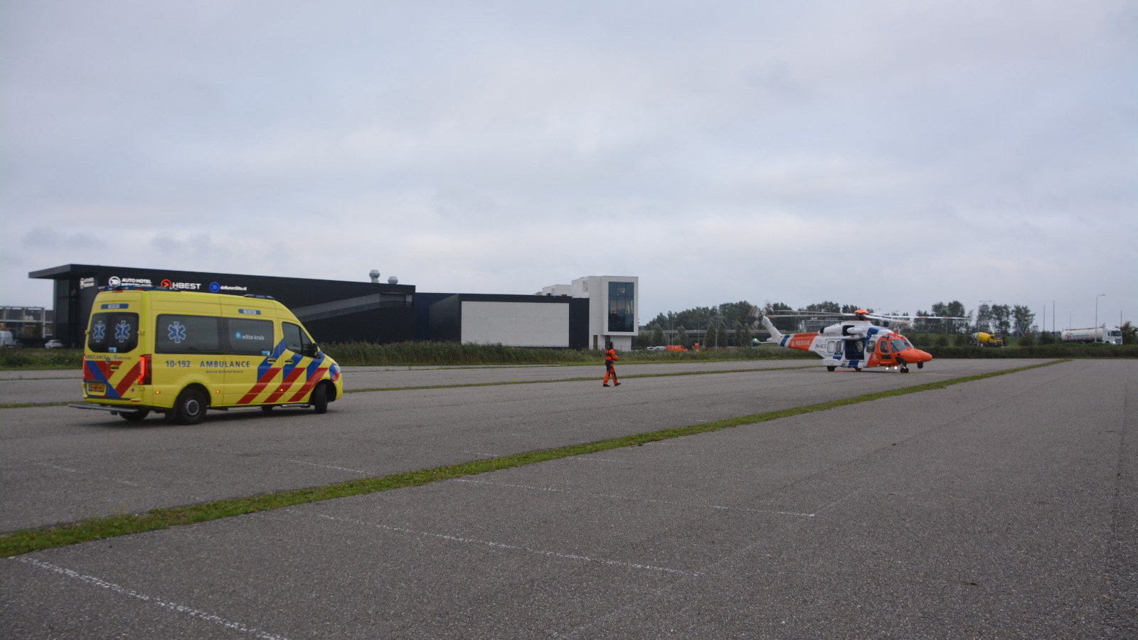 Geel-rode ambulance geparkeerd naast een helikopter in oranje en wit op een open terrein met gebouwen op de achtergrond.