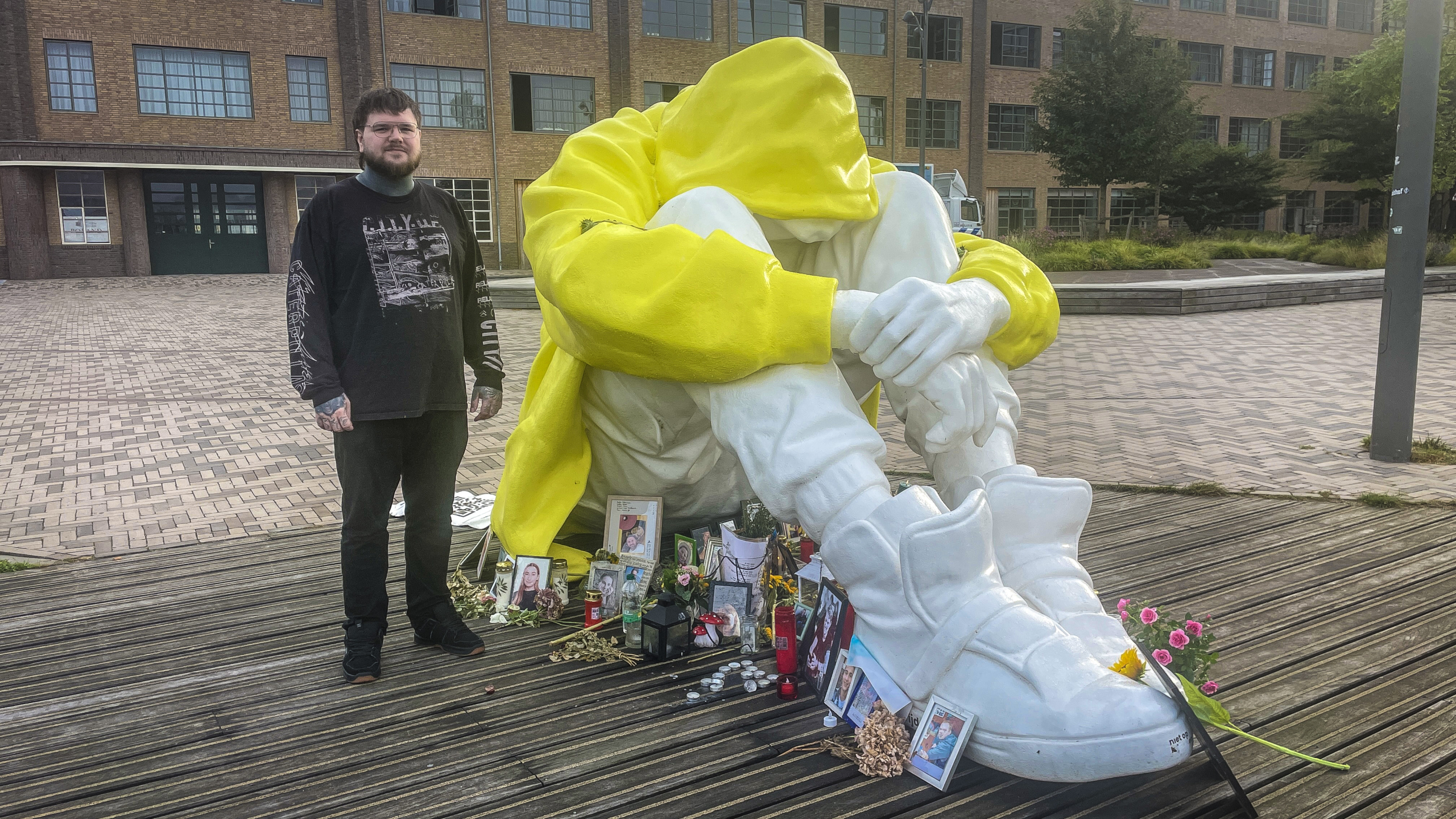 Man staat naast groot standbeeld van een persoon met gele hoodie en witte kleding, met verschillende herdenkingsitems zoals foto's en bloemen aan de voeten van het standbeeld, in een stedelijke omgeving met bakstenen gebouwen.