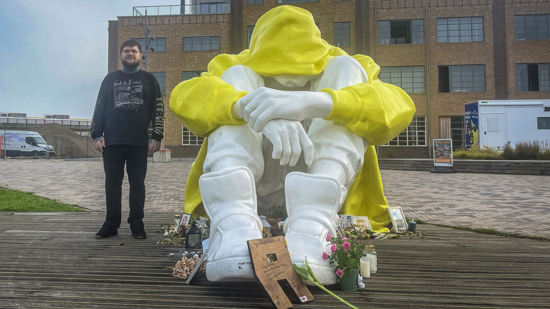 Beeld van een zittende figuur met gele capuchon op een plein, gefotografeerd naast een man.