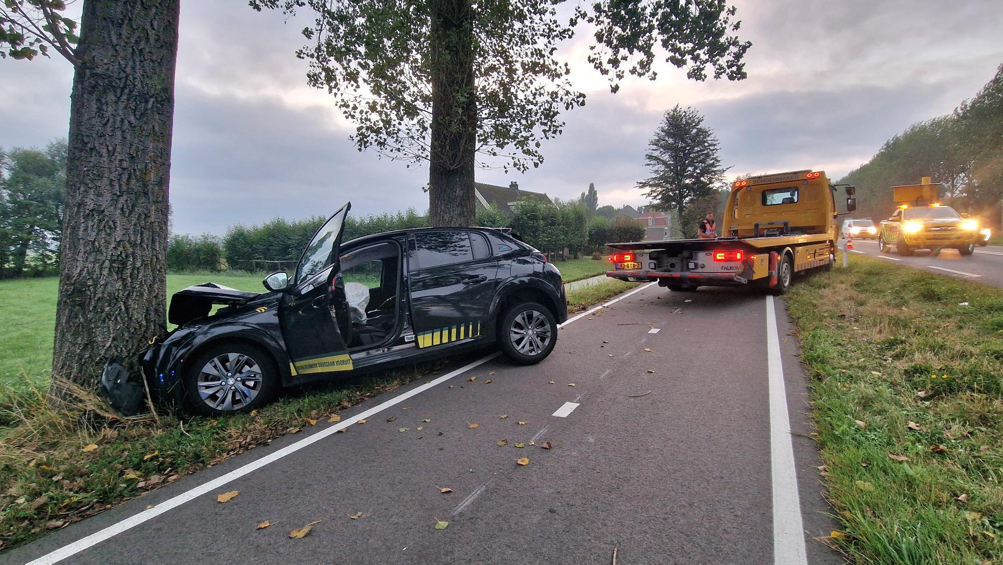 Auto gebotst tegen boom langs een weg, met een takelwagen en een ander voertuig ter plaatse.