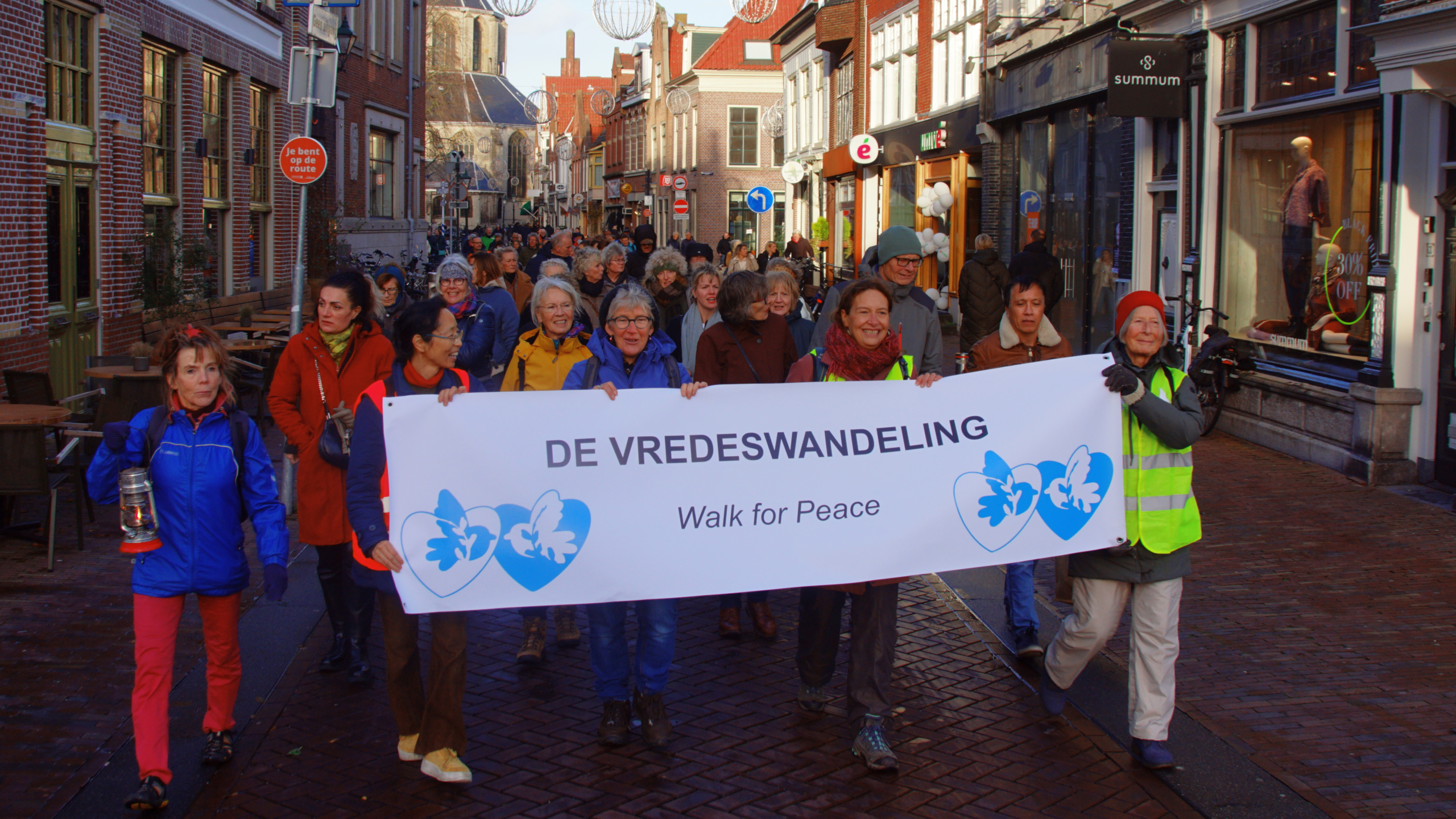 Groep mensen neemt deel aan de vredeswandeling in een stedelijke omgeving, met een spandoek waarop "DE VREDESWANDELING - Walk for Peace" staat.