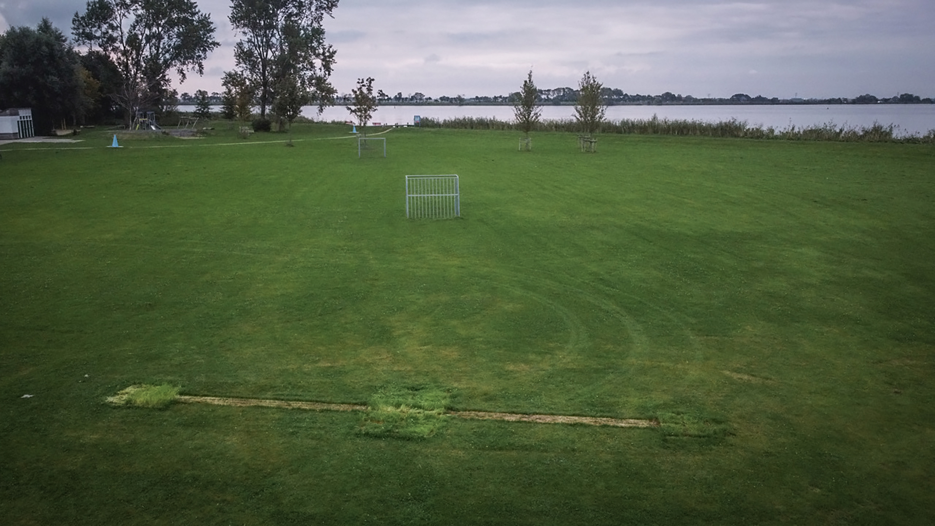 Groot open grasveld met metalen doelpalen, bomen op de achtergrond en een meer op een bewolkte dag.