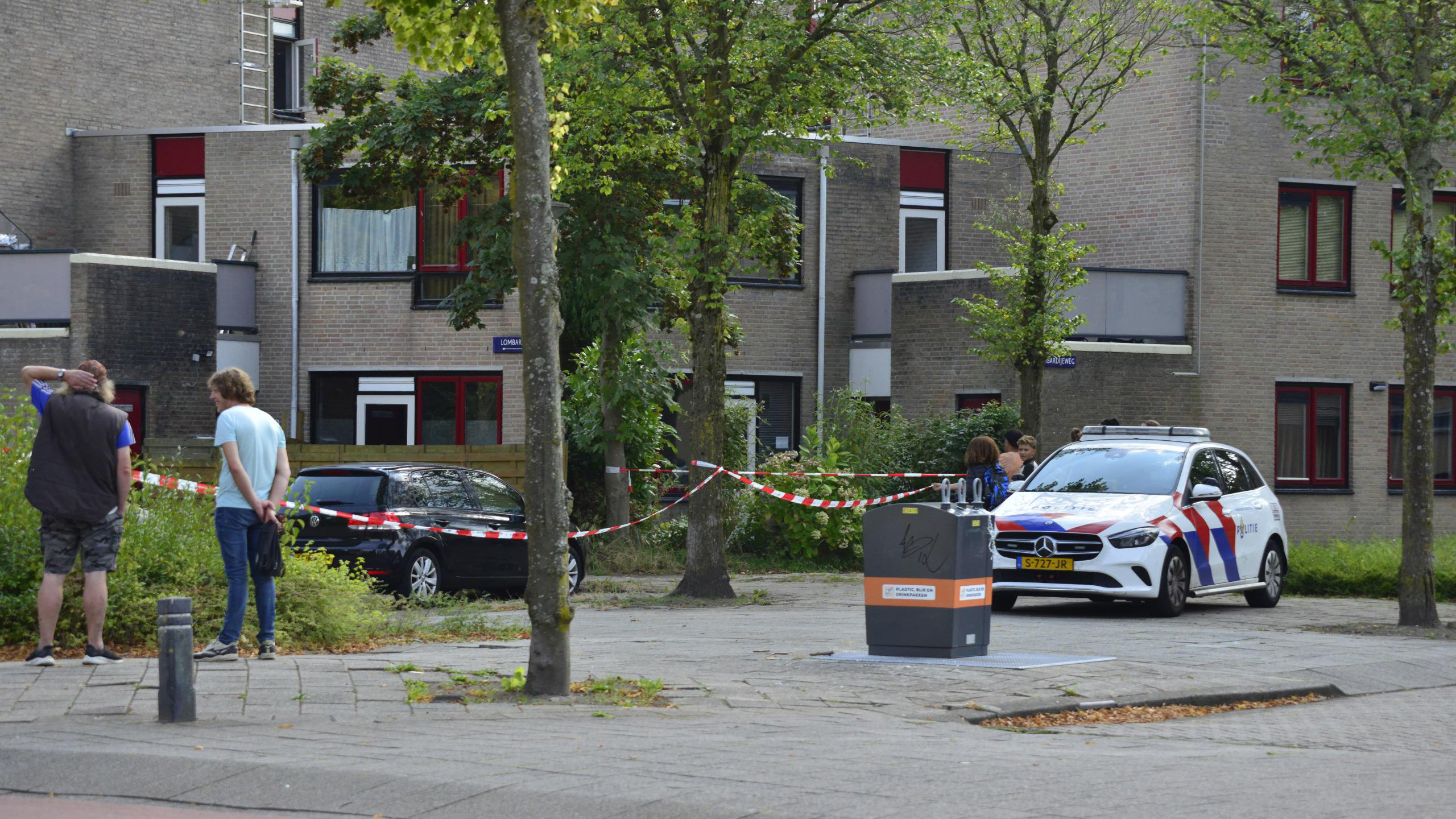 Politieauto en mensen bij een afgezette plaats in een woonwijk met bakstenen huizen en bomen.