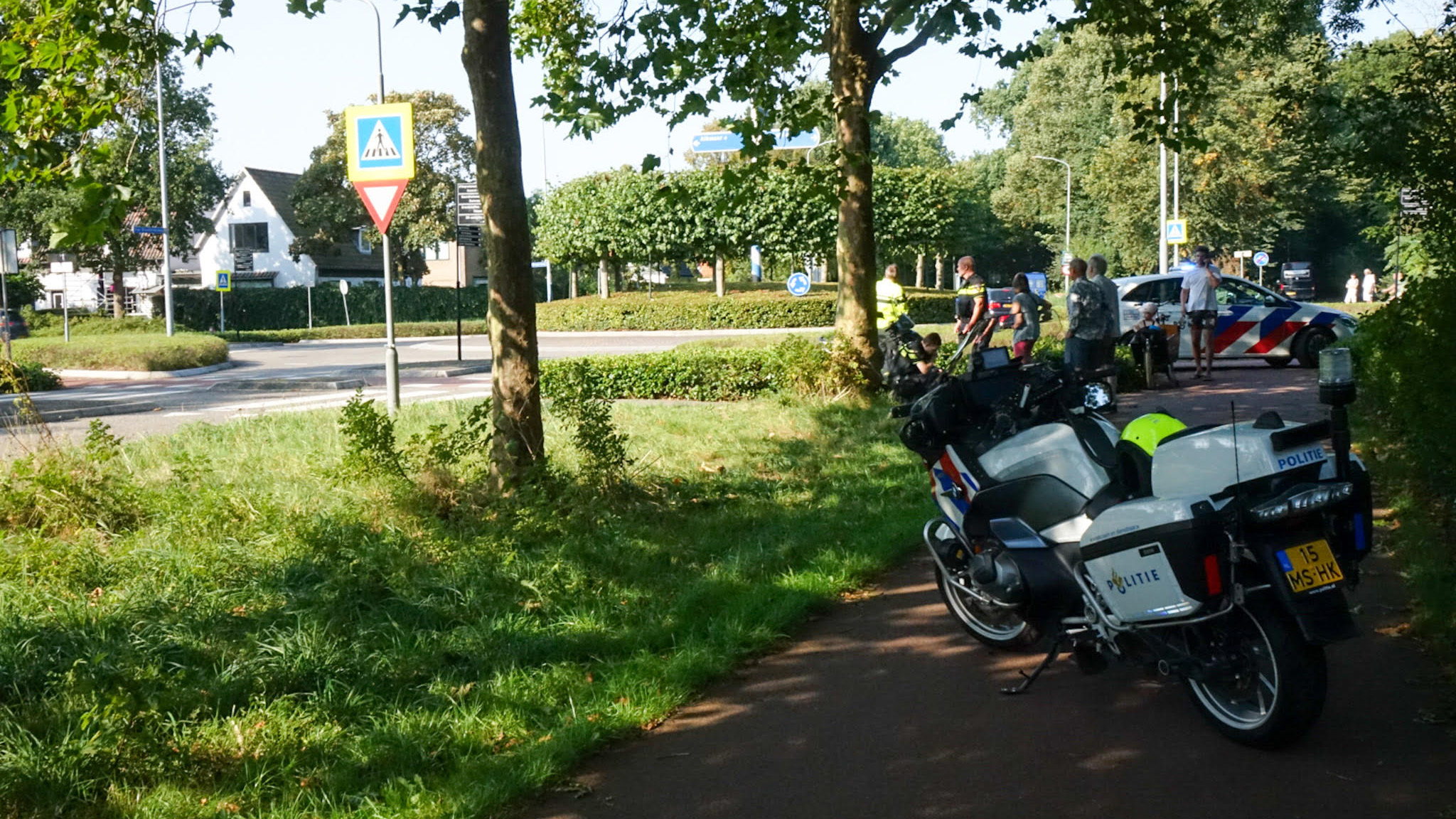 Twee politiefietsen en een politieauto bij een kruispunt, omgeven door bomen en gras, met meerdere mensen en agenten op de achtergrond.