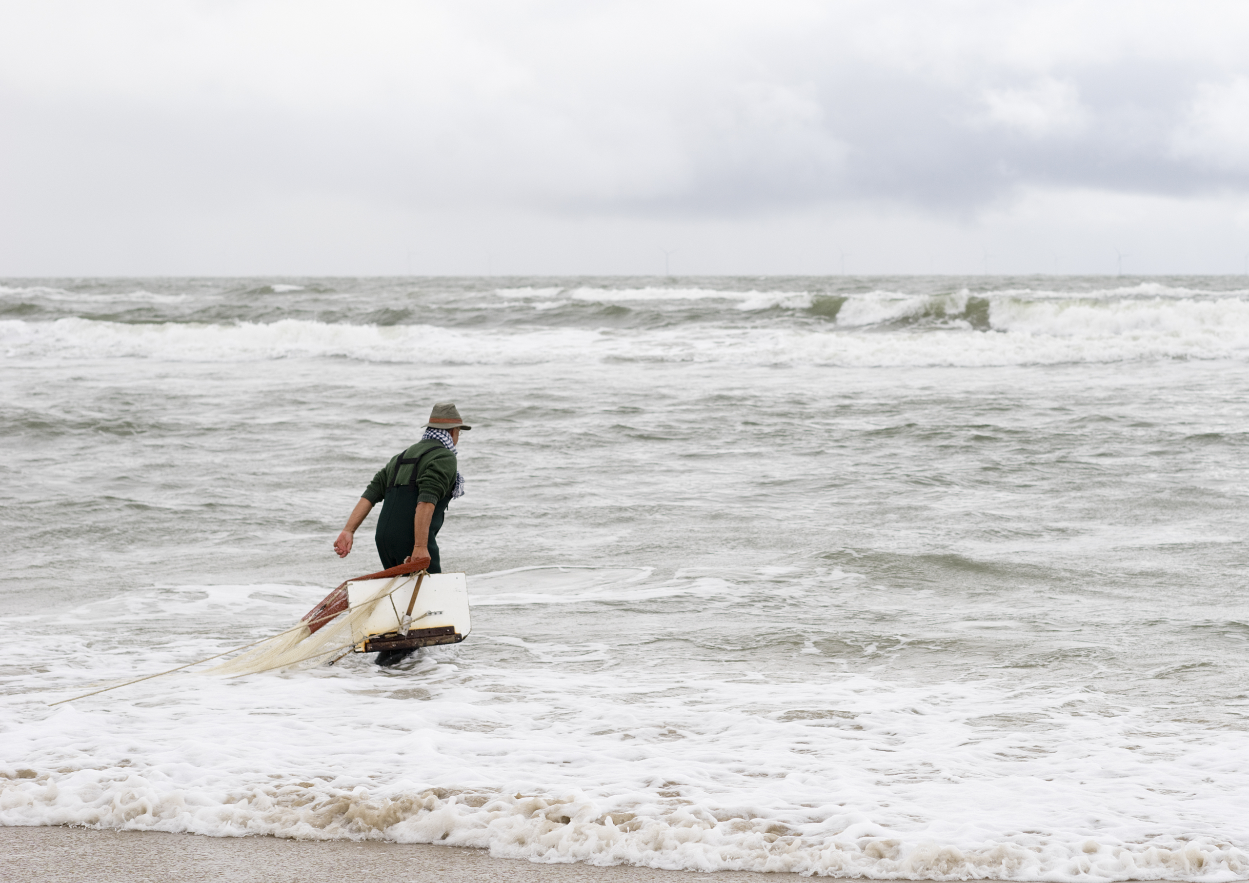 Persoon die met een visnet de zee inloopt op een bewolkte dag.