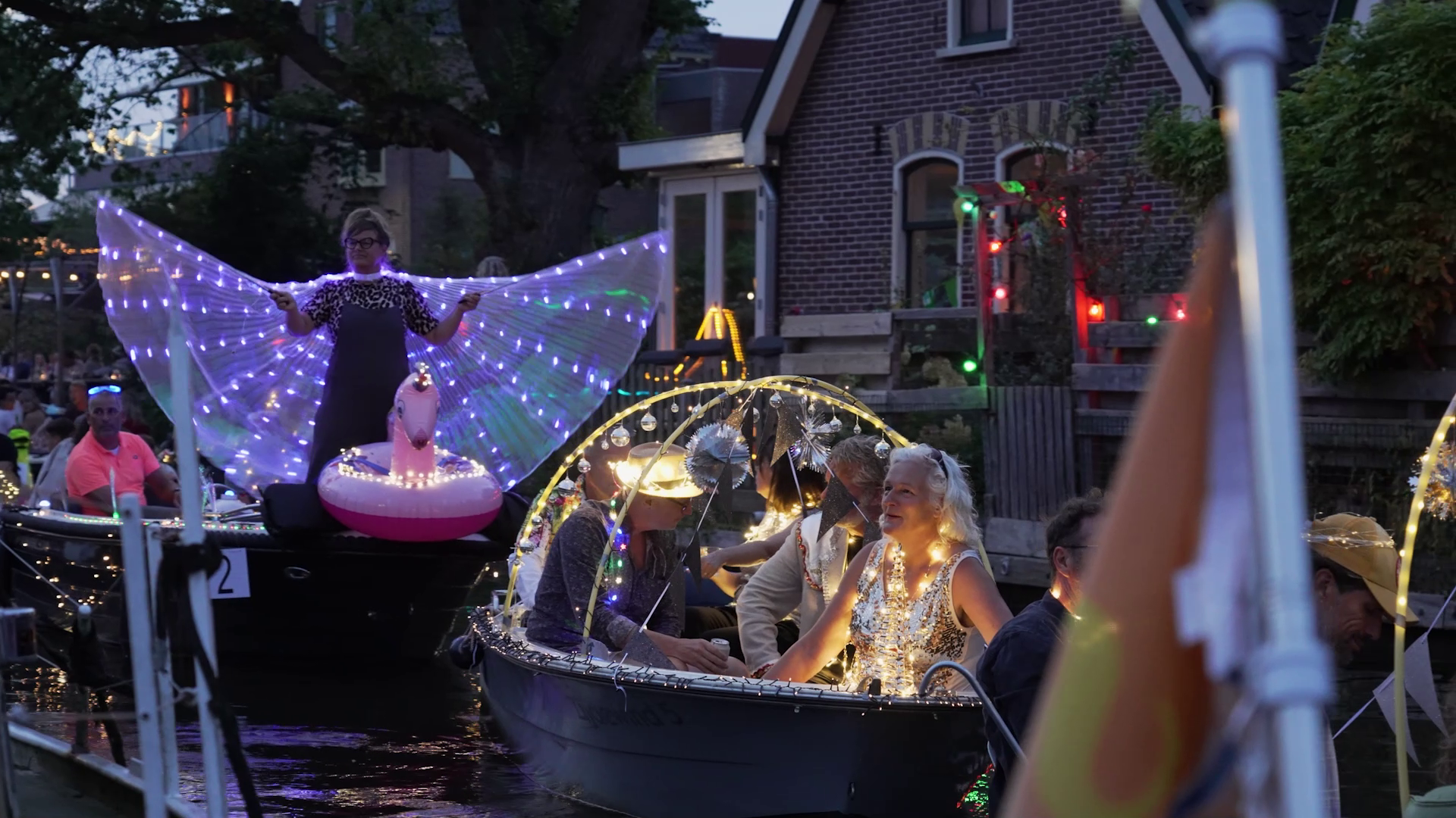 Boten versierd met kleurrijke LED-verlichting tijdens een avondparade op een gracht, met mensen in opvallende kostuums.