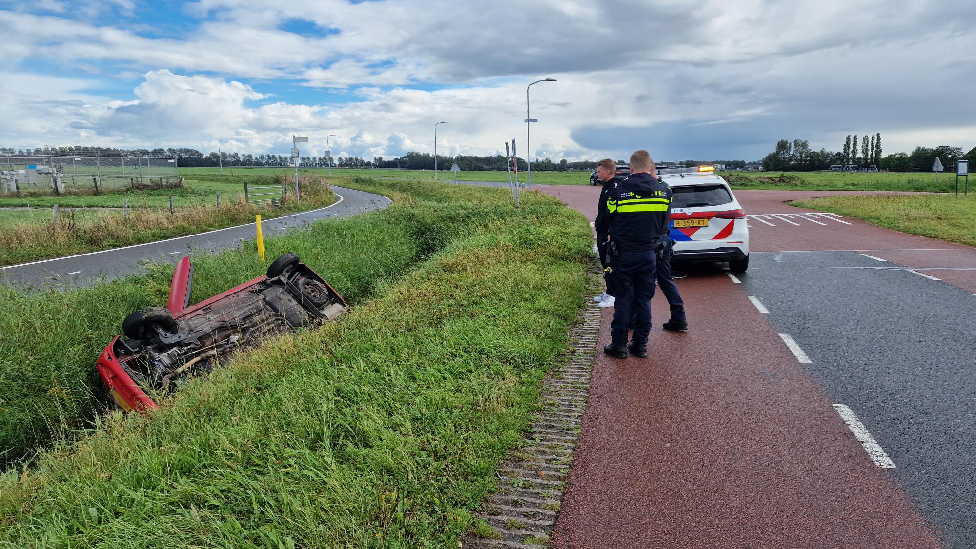 Auto op zijn kop in de berm naast een weg, met politieagenten en een politieauto.