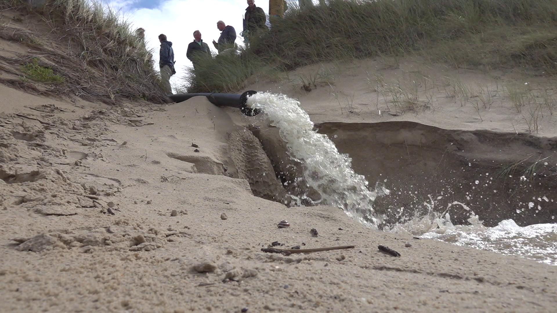 Pijp die water op zandige grond stort met mensen op een duin op de achtergrond.