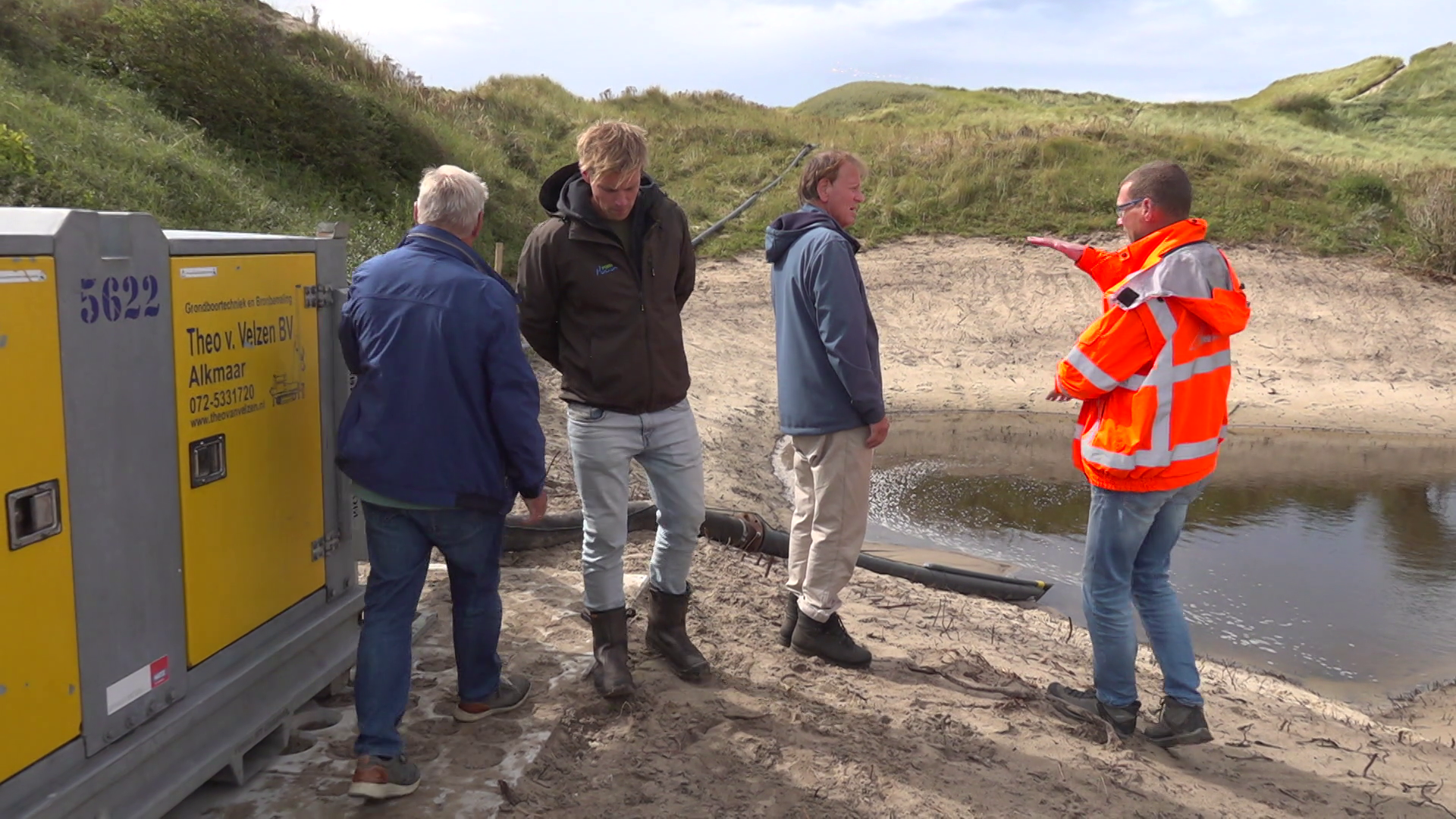 Vier mannen staan bij een bouwplaats naast een zandduin, met een gele machine links en een kleine waterplas rechts. Eén man draagt een fel oranje veiligheidsjas.