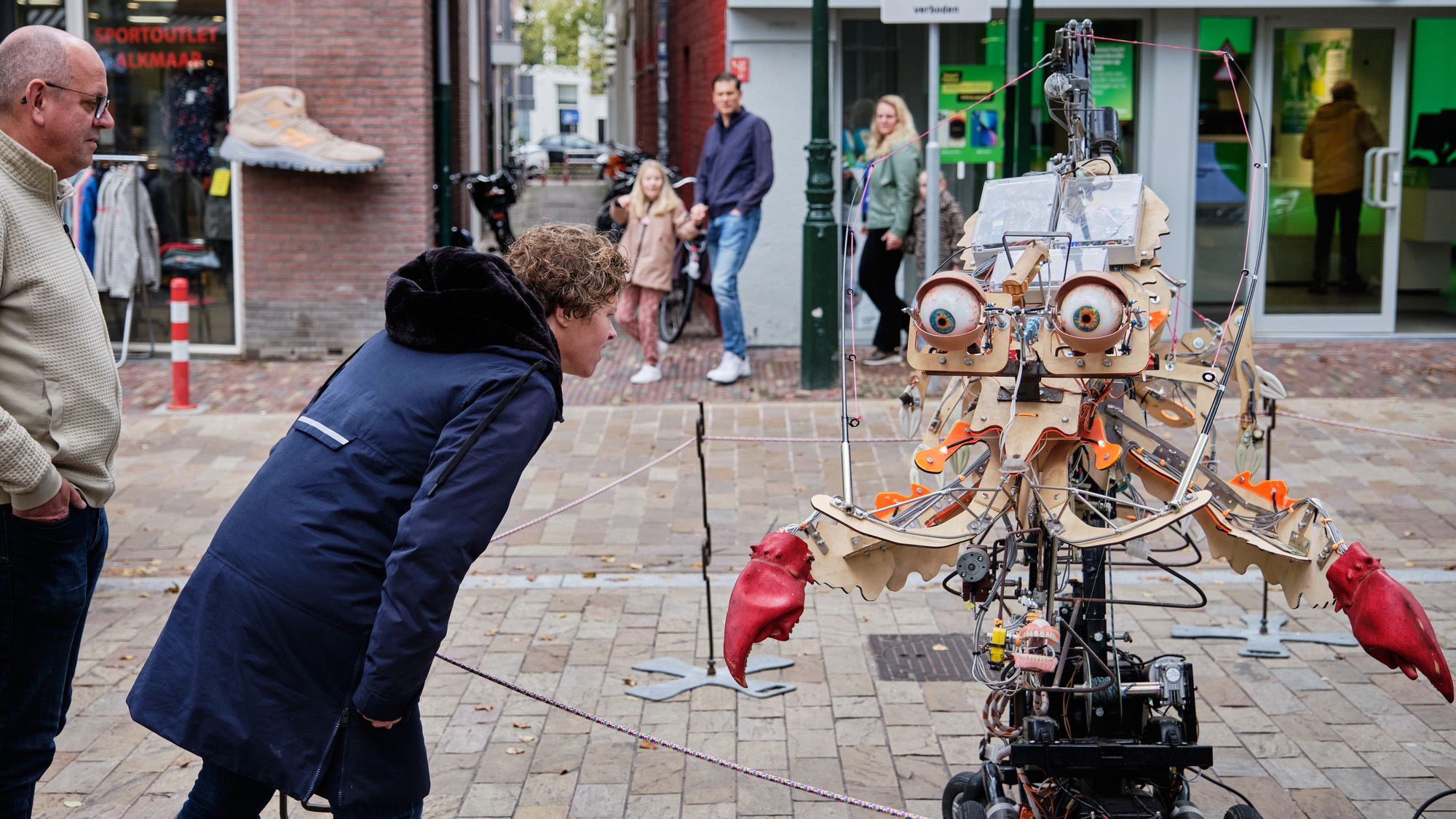 Een man en een vrouw buigen zich voorover om een geavanceerde robot met grote ogen en rode klauwen op een straat te bekijken, terwijl andere mensen in de achtergrond staan en toekijken.