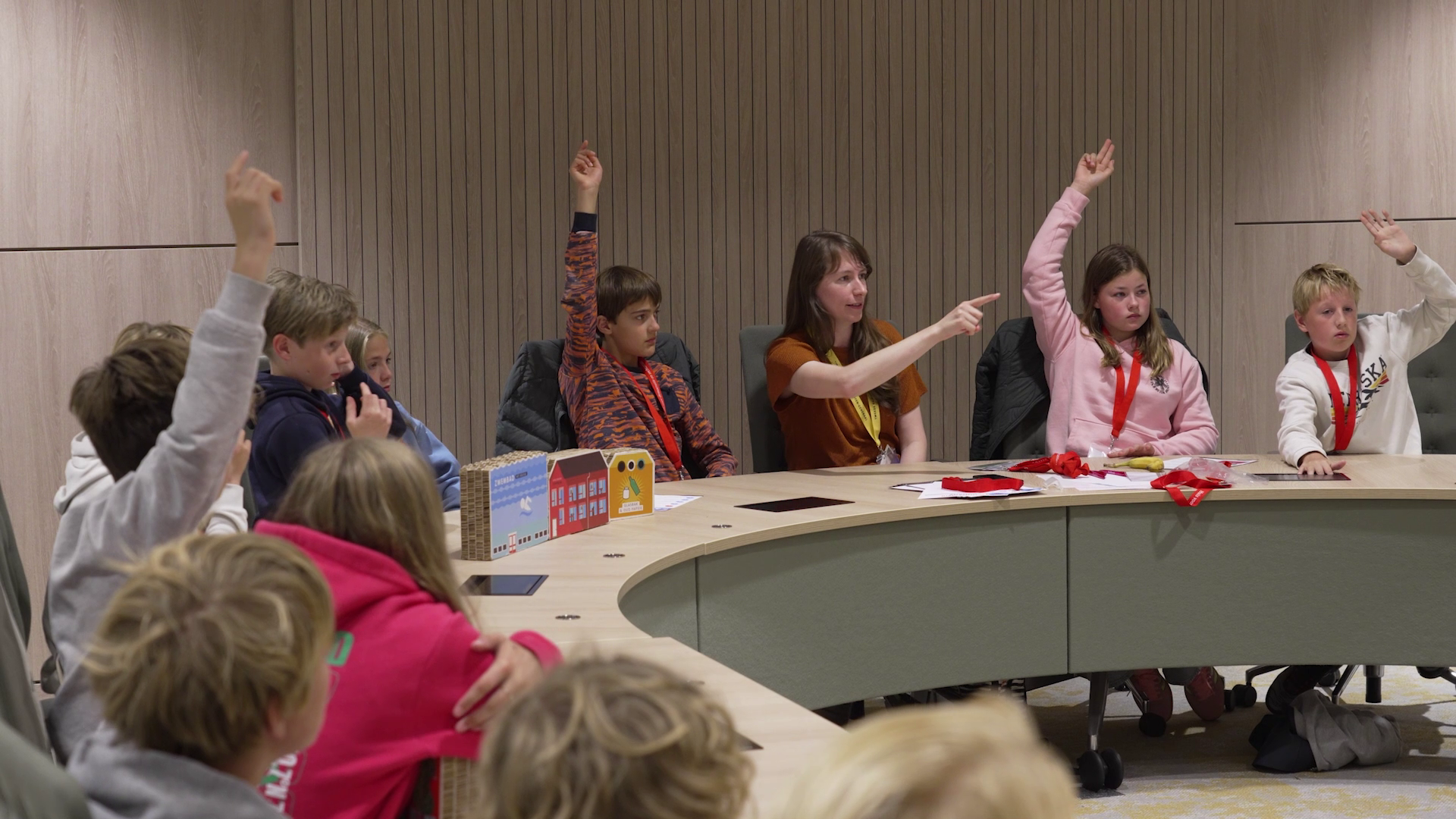 Kinderen zitten rond een tafel in een vergaderruimte met enkele kinderen die hun hand opsteken.