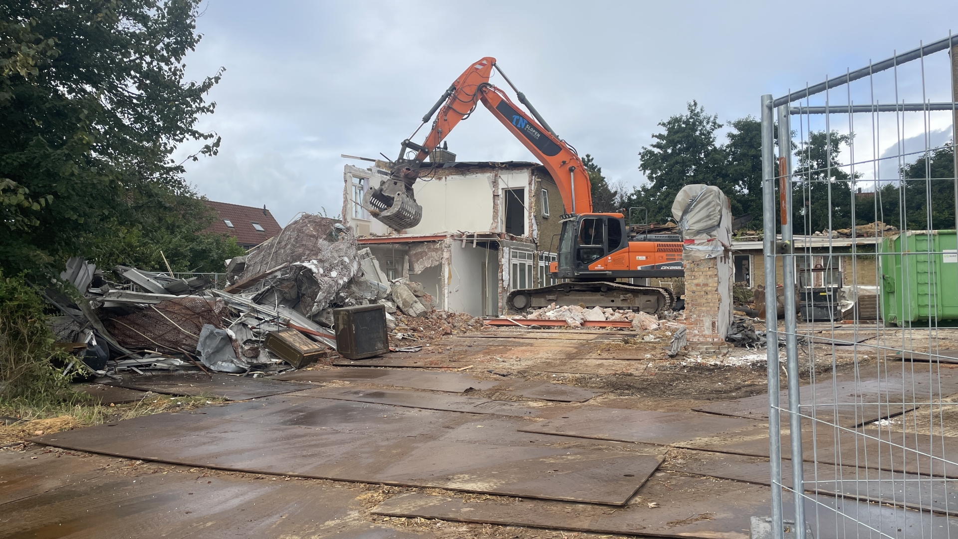 Graafmachine sloopt een gebouw omringd door puin en een hek.