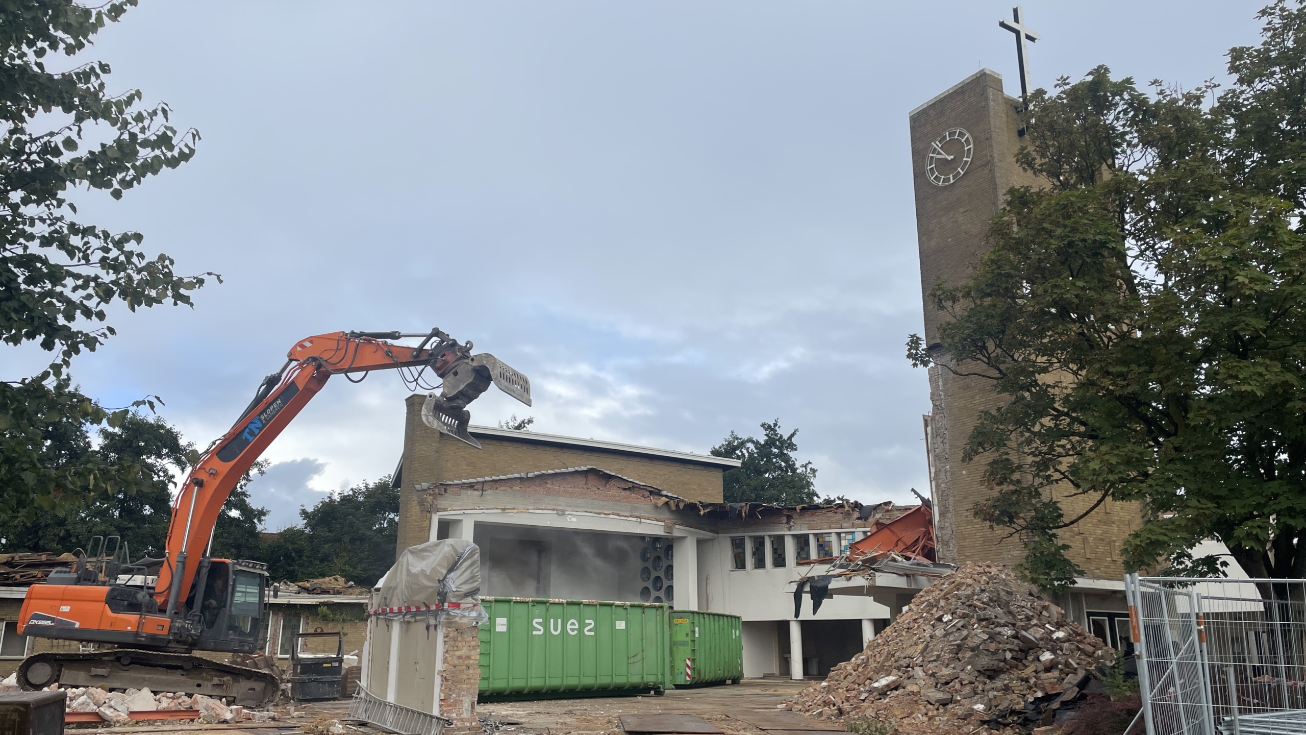 Een bouwplaats waarop een kerk met een klokkentoren wordt gesloopt door een oranje graafmachine, omgeven door puin en een groene container.