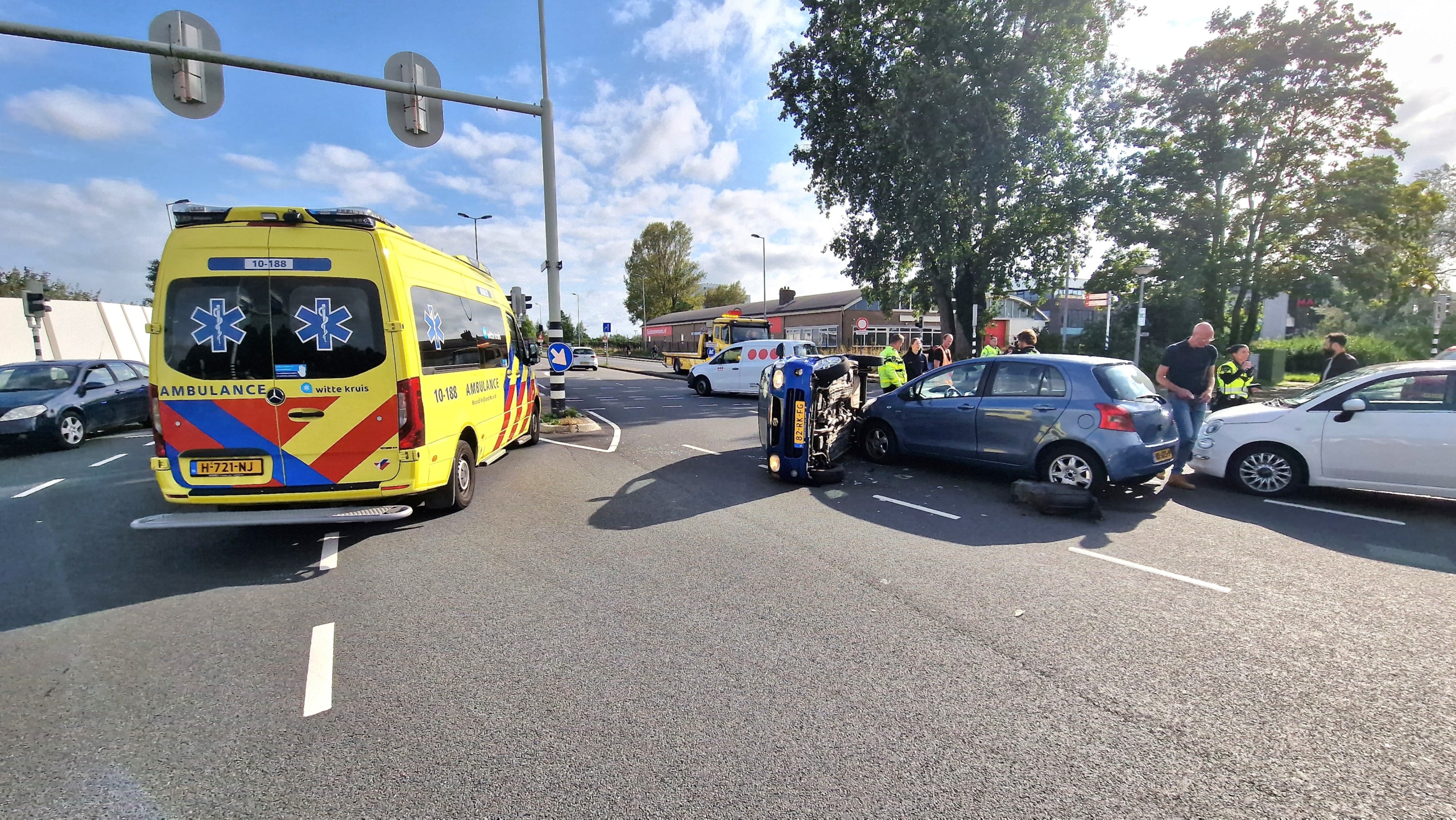 Verkeersongeluk met een gekantelde auto, een blauwe auto en een witte auto op een kruispunt, met een ambulance ter plaatse en omstanders en hulpverleners aanwezig.