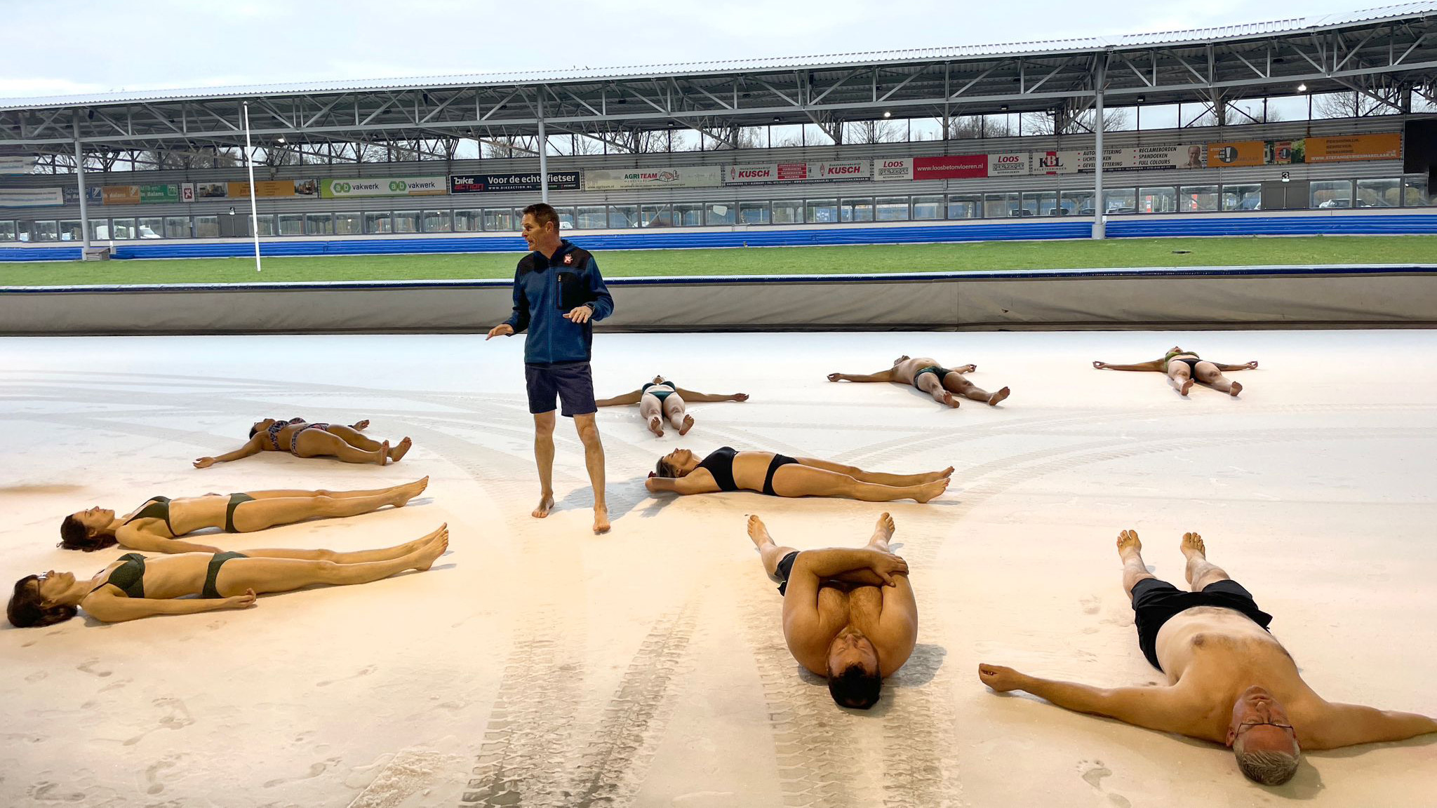 Mensen in zwemkleding mediteren op een besneeuwd oppervlak in een sportstadion, met een instructeur in het midden.