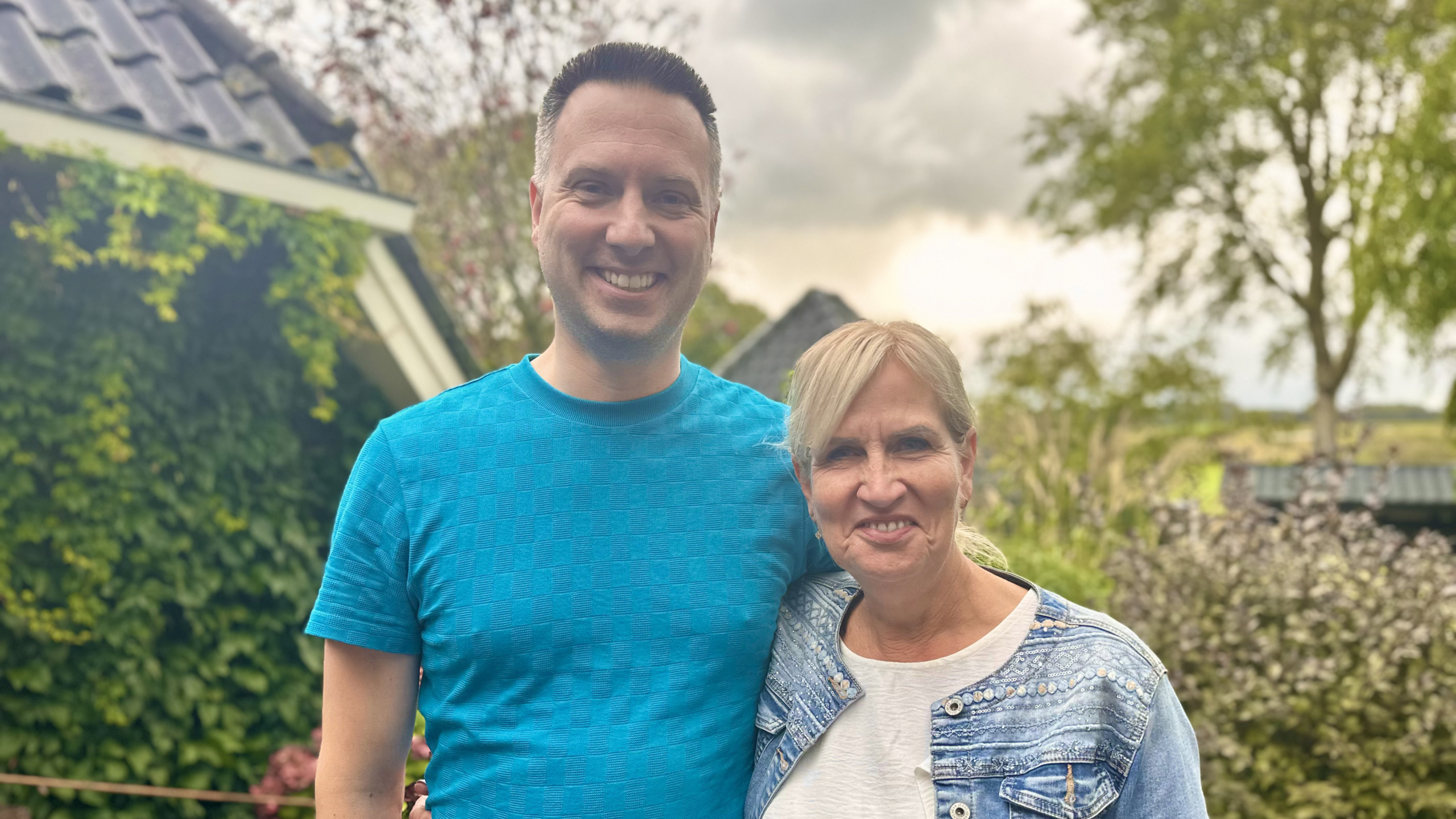 Man en vrouw poseren buiten in een tuin, beiden glimlachend naar de camera.