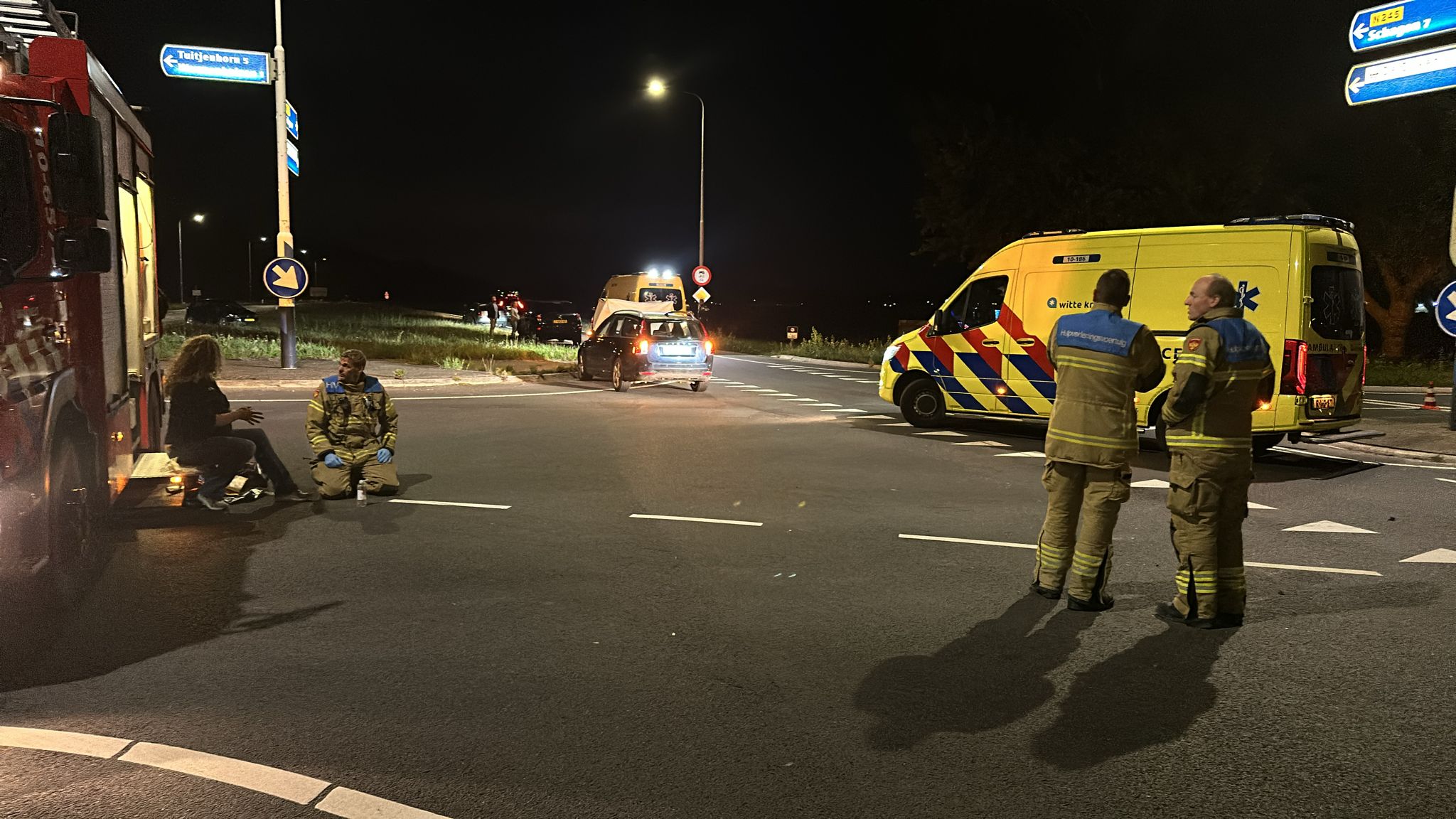 Brandweerlieden en een vrouw bij een ongeval met meerdere voertuigen 's nachts met een ambulance op de achtergrond.