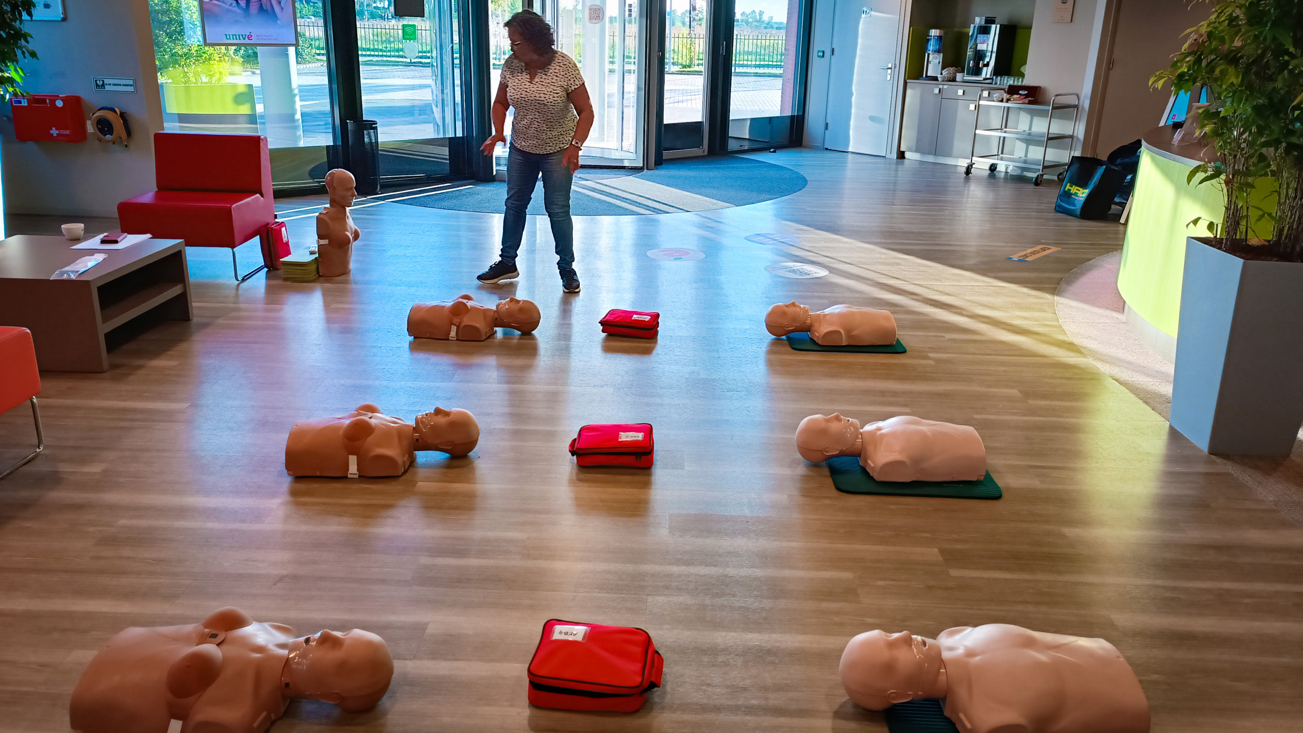 Een vrouw begeleidt een reanimatietraining met meerdere oefenpoppen en EHBO-tassen in een ruime, goed verlichte kamer.